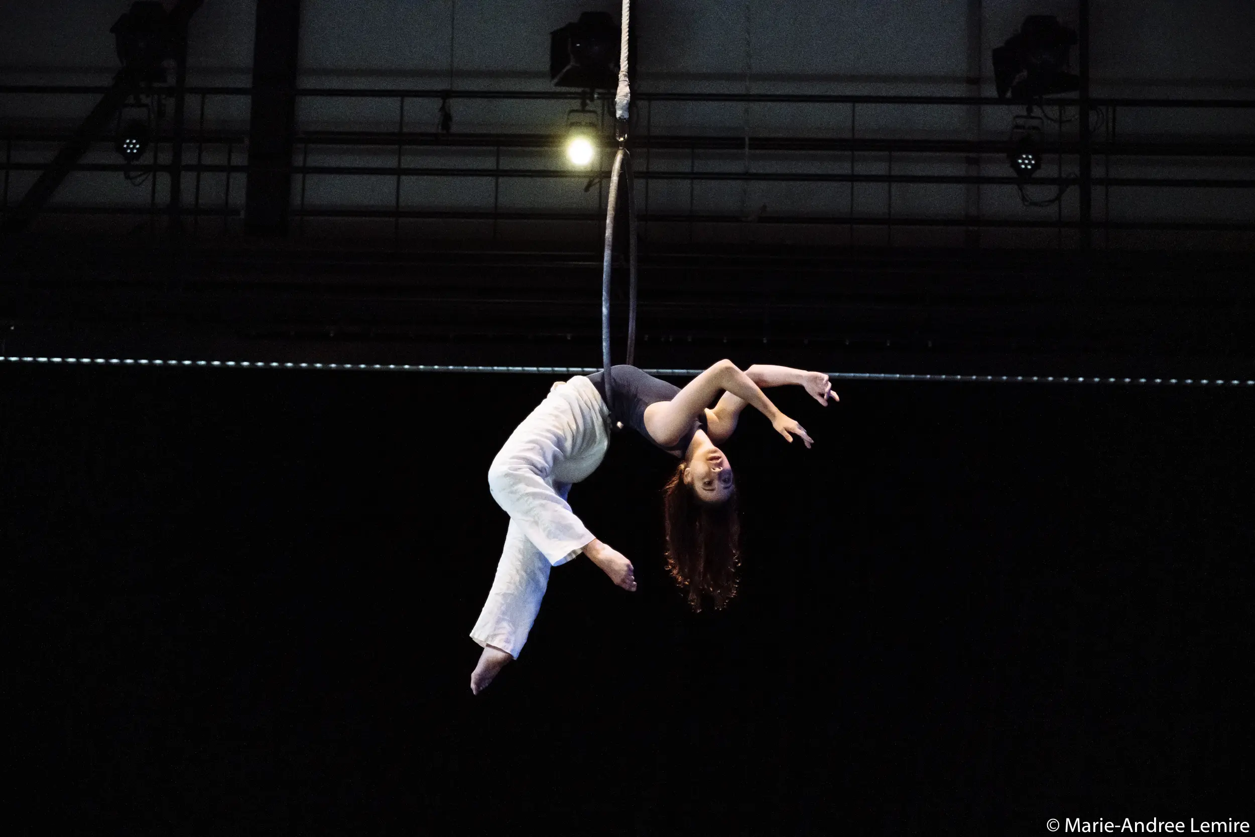 L'artiste aérienne Michelle Sargent est suspendue à l'envers sur un cerceau, vêtue d'un pantalon blanc et d'un haut gris, les bras gracieusement étendus sur un fond sombre.