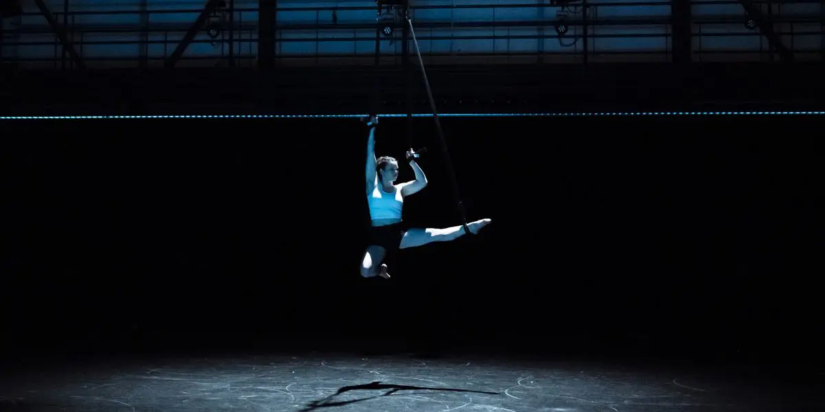 Clara Laurent exécute un saut en l'air sur un cerceau aérien dans un théâtre sombre et dramatique, éclairé par un projecteur depuis le haut. La scène et l'arrière-plan sont faiblement éclairés, ce qui met en valeur sa pose saisissante et l'ombre qu'elle projette.