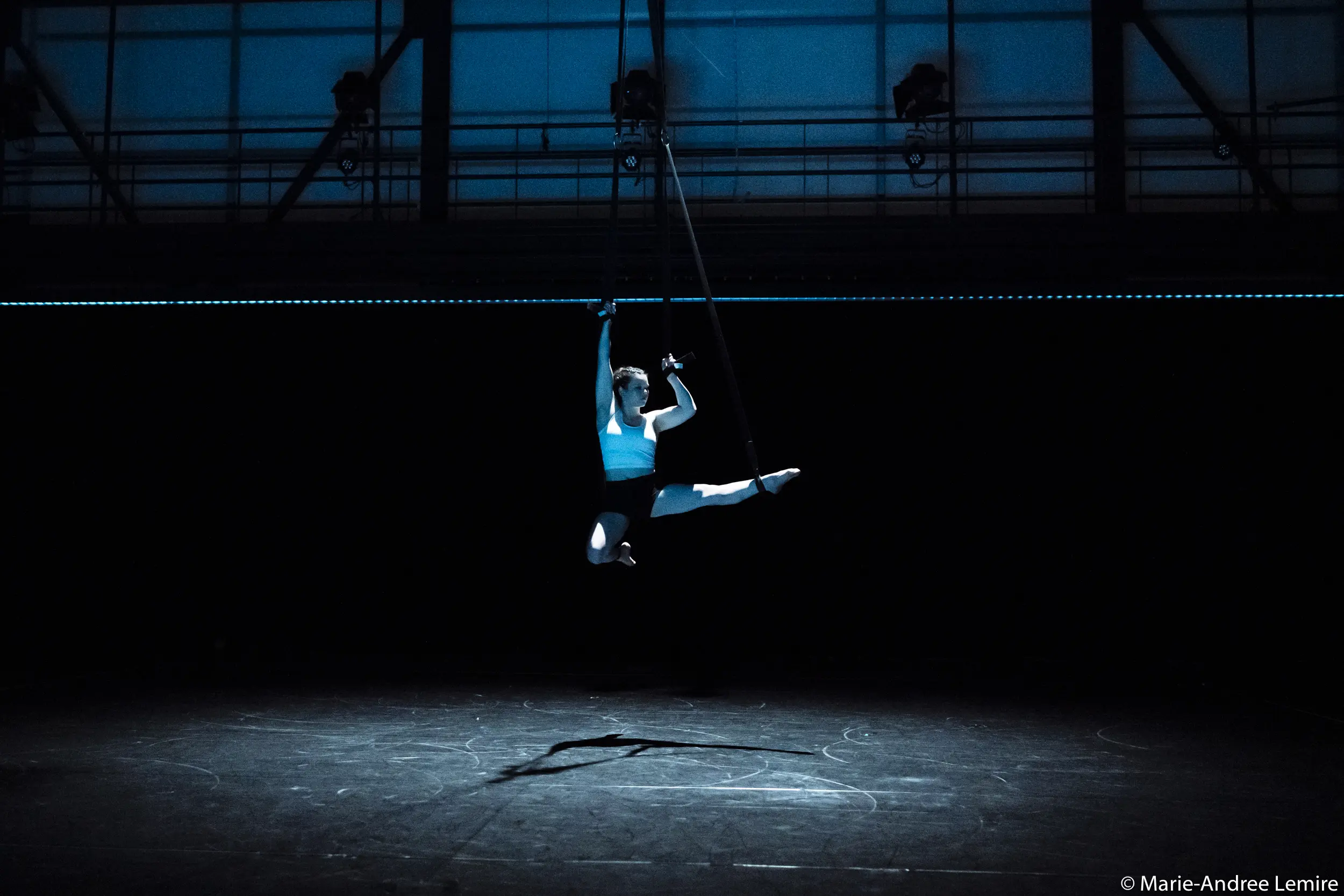 Clara Laurent exécute un saut en l'air sur un cerceau aérien dans un théâtre sombre et dramatique, éclairé par un projecteur depuis le haut. La scène et l'arrière-plan sont faiblement éclairés, ce qui met en valeur sa pose saisissante et l'ombre qu'elle projette.
