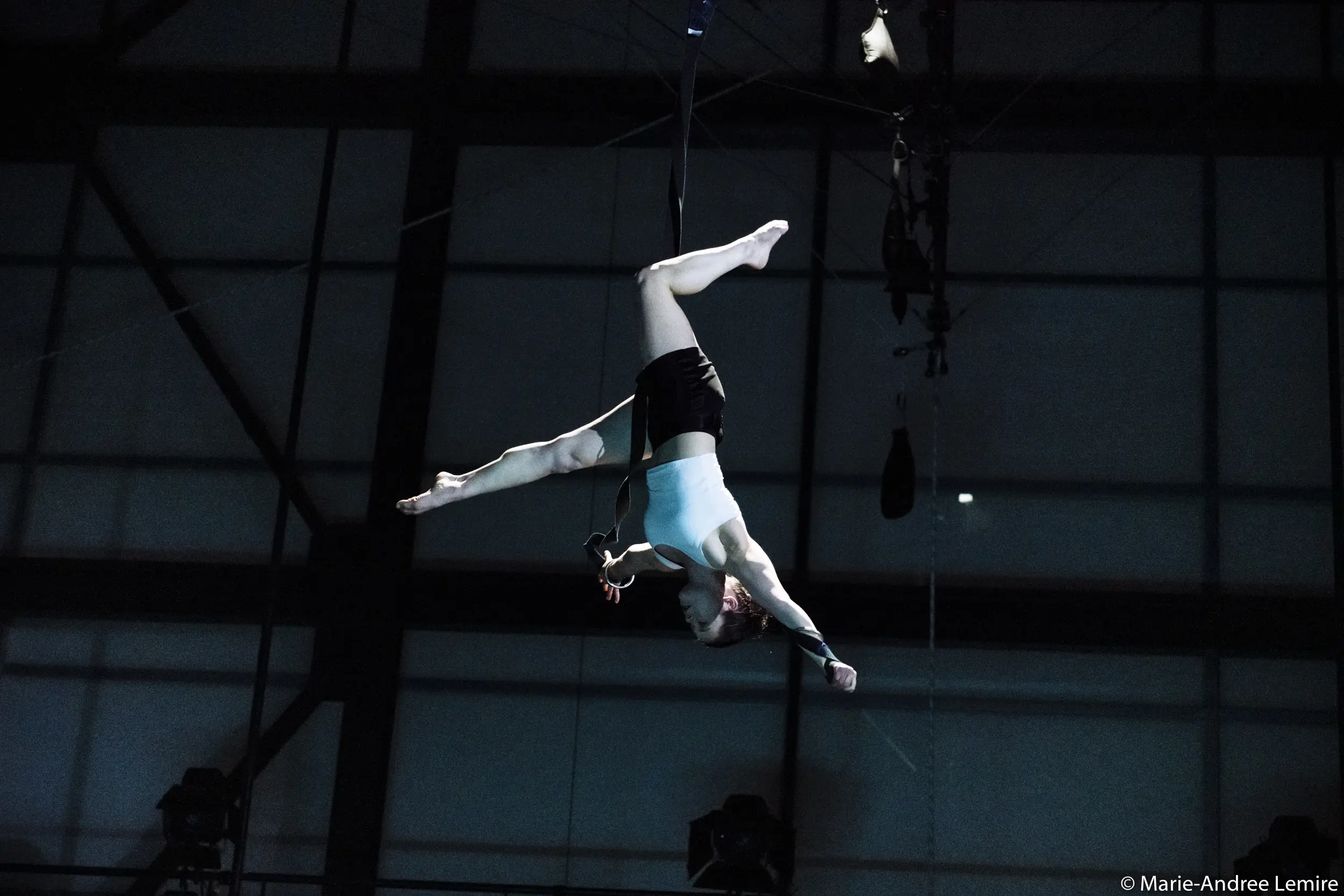 L'artiste aérienne Clara Laurent exécute un grand écart inversé, suspendue à des sangles dans une salle intérieure faiblement éclairée, avec des poutres métalliques visibles à l'arrière-plan.