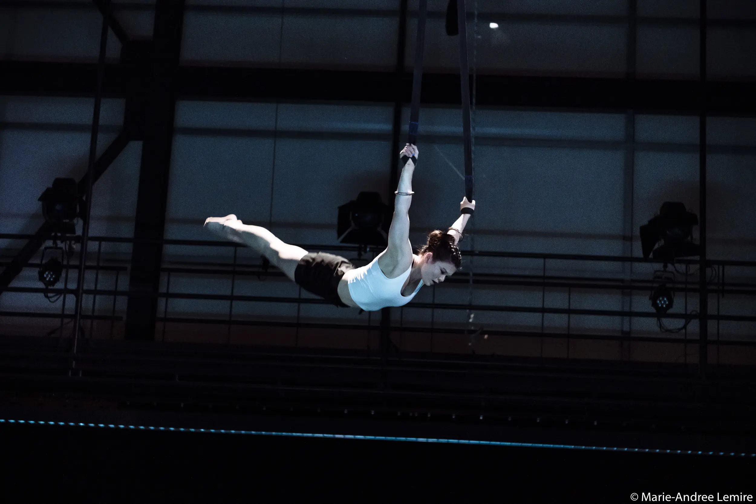 Clara Laurent, vêtue d'un débardeur blanc et d'un short noir, exécute un numéro aérien, s'accrochant à des sangles suspendues et étendant son corps à l'horizontale dans un espace intérieur faiblement éclairé.