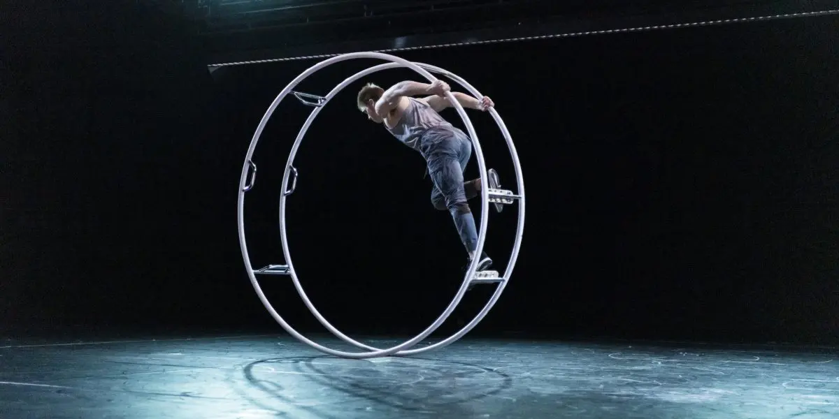 Elijah Kirk se tient en équilibre dans une grande roue de cirque en métal, appelée roue Cyr, sur une scène faiblement éclairée par des projecteurs.