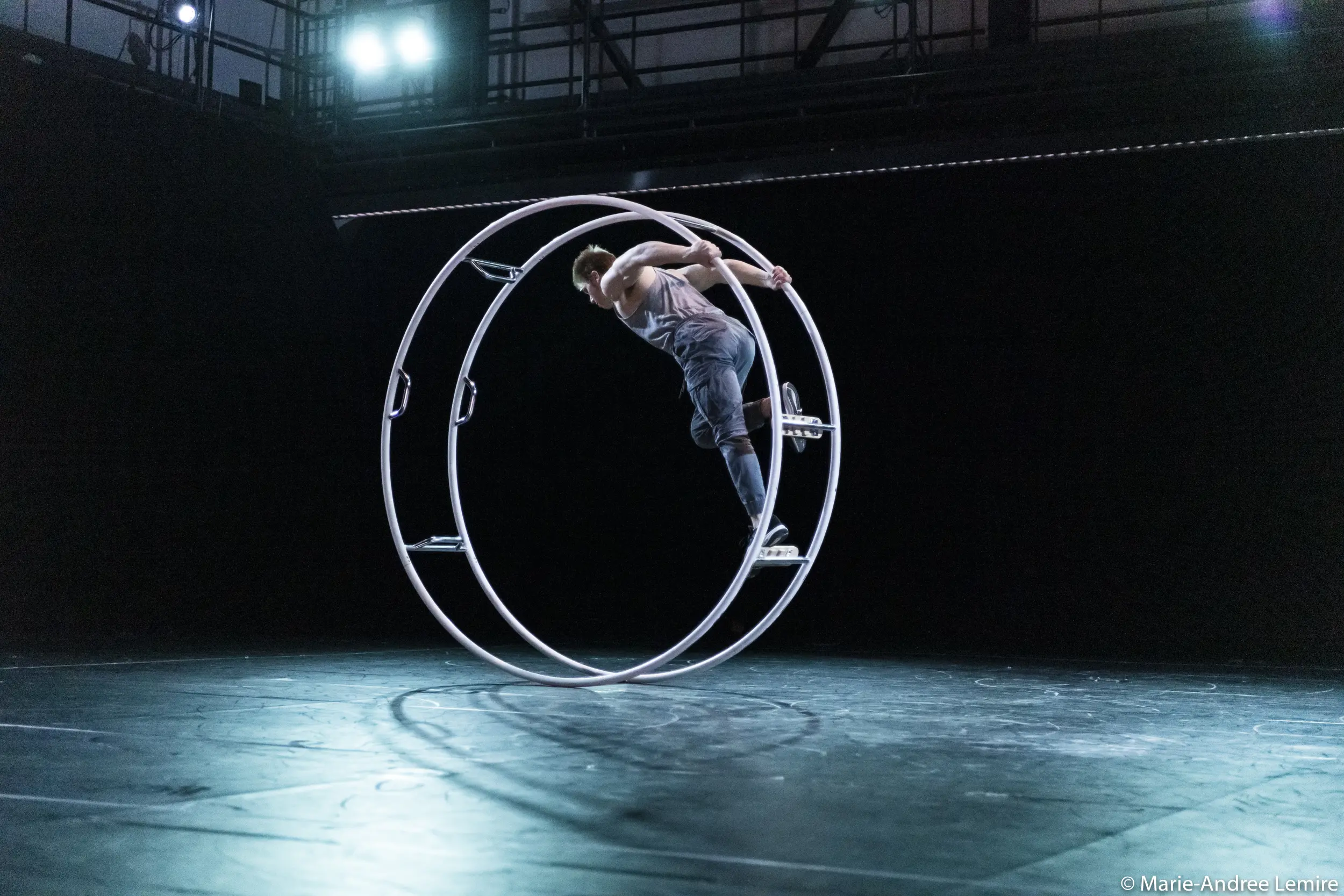 Elijah Kirk se tient en équilibre dans une grande roue de cirque en métal, appelée roue Cyr, sur une scène faiblement éclairée par des projecteurs.