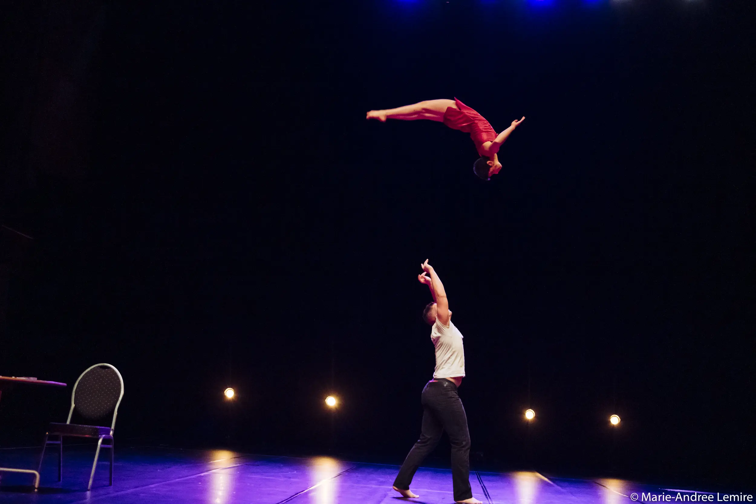 Un artiste se tient sur scène, les bras levés, tandis que Michael Carter et Frida Velasco éblouissent la foule - Frida s'envolant à l'envers dans une tenue rouge en hauteur. Les lumières de la scène et une chaise créent un décor dramatique à l'arrière-plan.