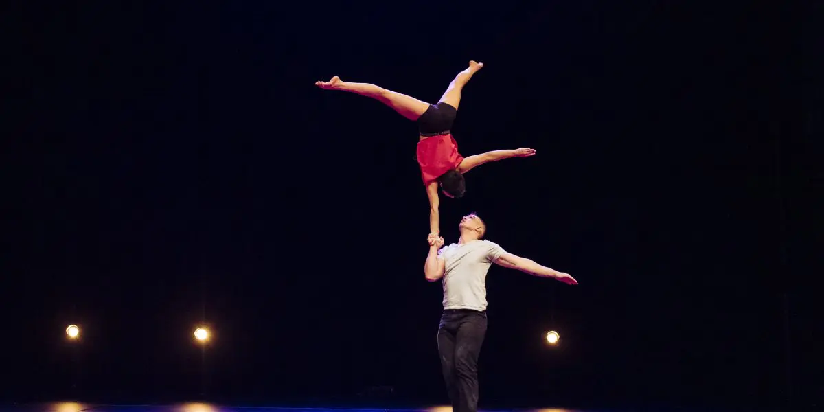 Deux acrobates, Michael Carter et Frida Velasco, se produisent sur scène ; il se tient sur une jambe et équilibre Frida à l'envers dans une fente sur sa main, tandis que quatre lumières brillent à l'arrière-plan.