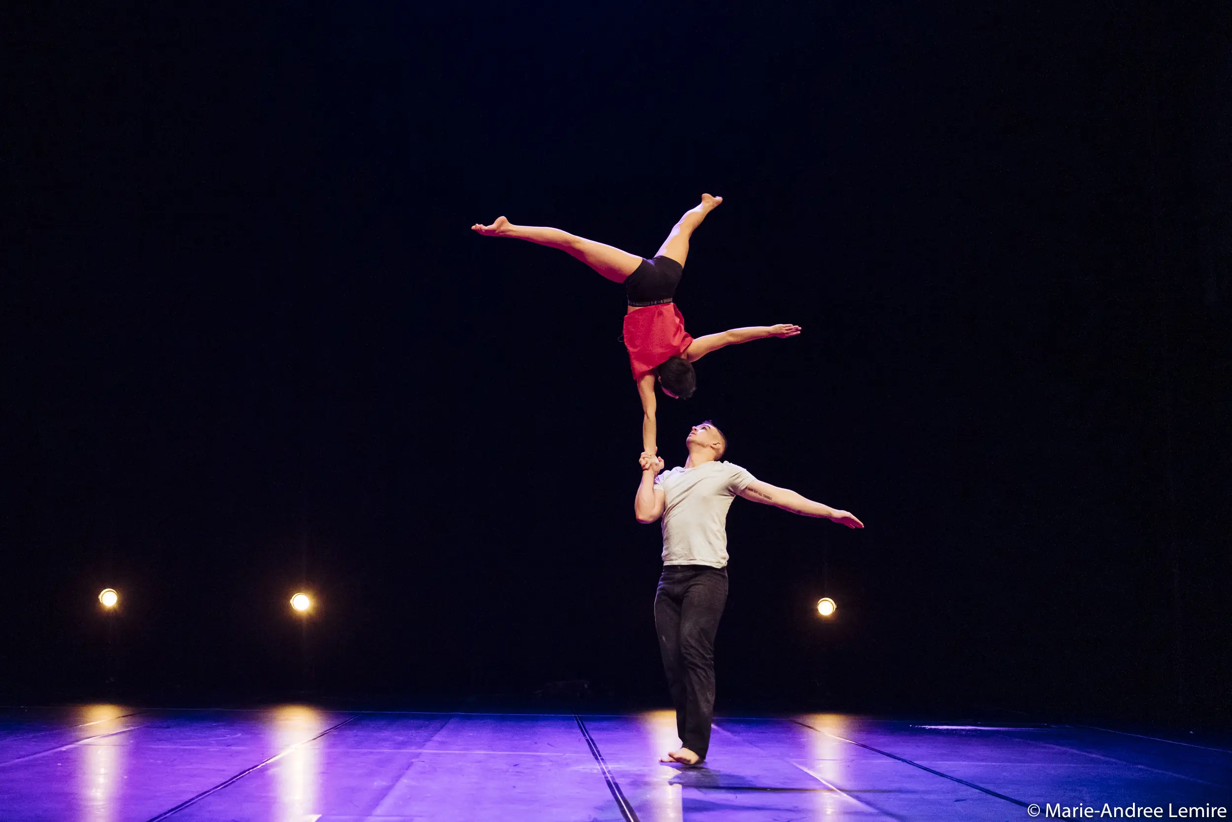 Deux acrobates, Michael Carter et Frida Velasco, se produisent sur scène ; il se tient sur une jambe et équilibre Frida à l'envers dans une fente sur sa main, tandis que quatre lumières brillent à l'arrière-plan.
