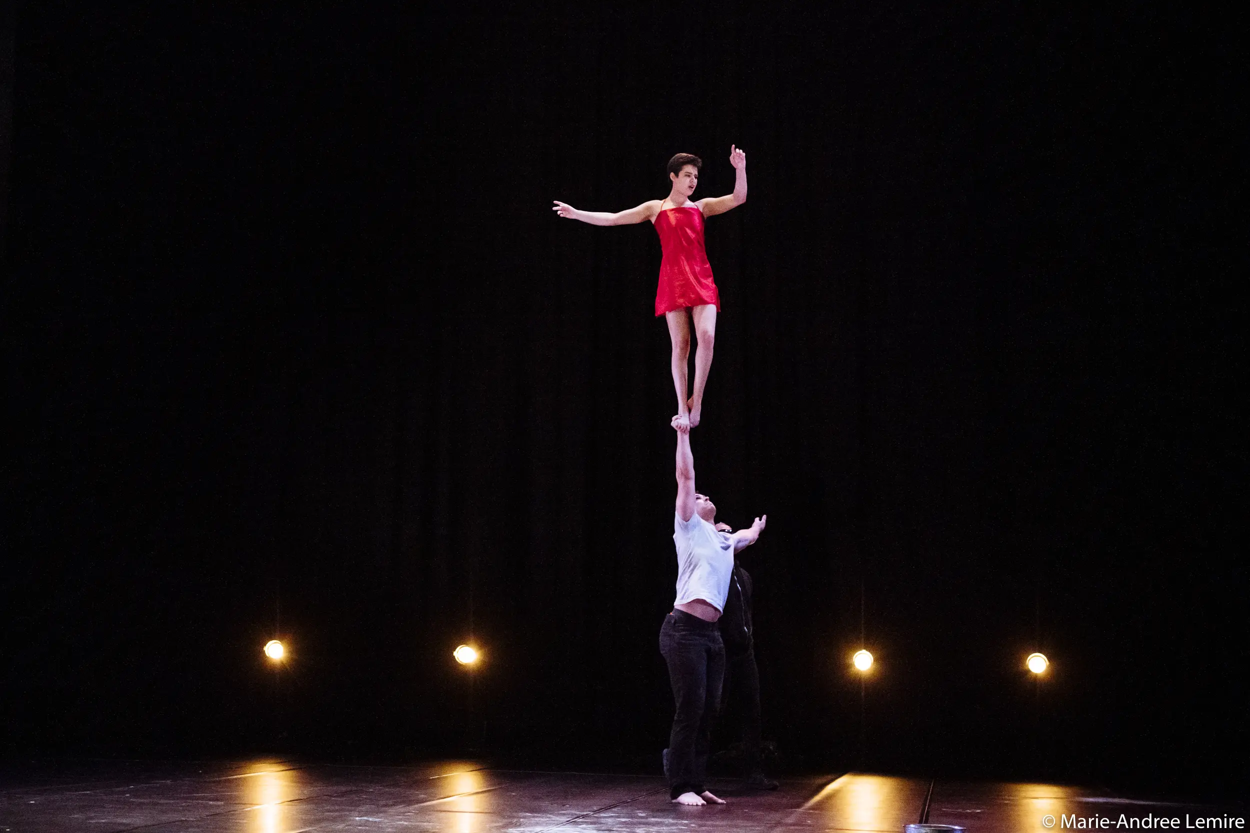 Michael Carter et Frida Velasco interprètent un homme qui tient en équilibre une femme vêtue d'une robe rouge au-dessus de sa tête avec un bras, lors d'une représentation sur une scène faiblement éclairée avec quatre projecteurs en arrière-plan.