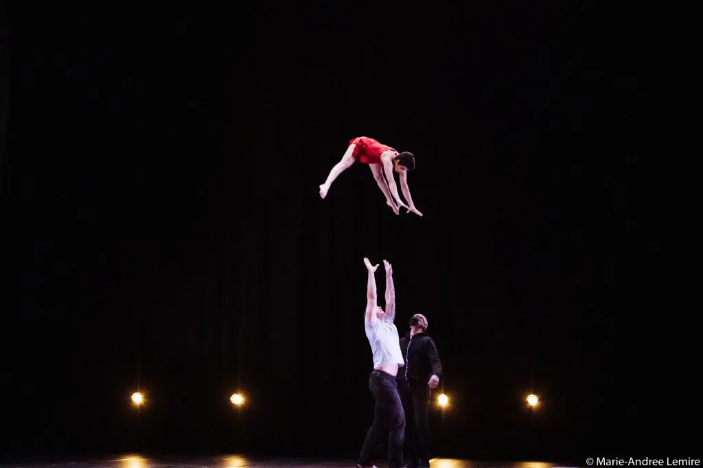 Une personne en rouge, peut-être Michael Carter et Frida Velasco, s'envole d'un bond au-dessus de deux artistes sur une scène sombre, dont l'un a les bras levés. Des lumières vives éclairent le fond de la scène.