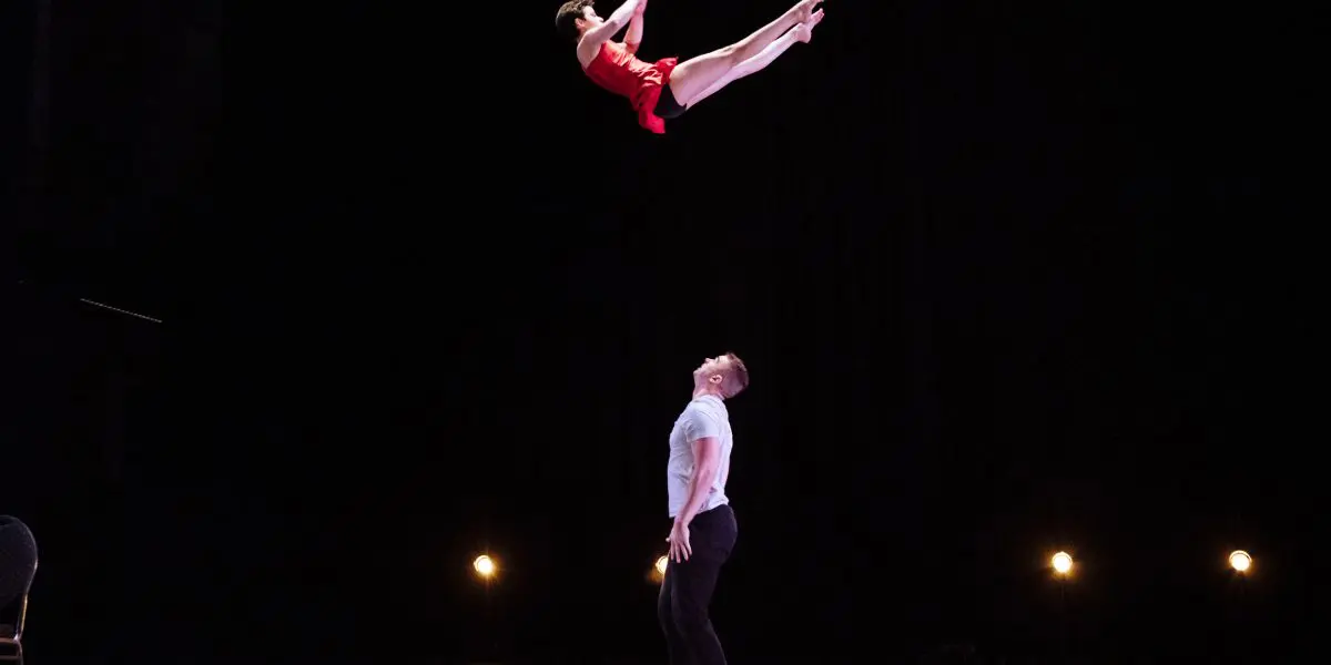 Un artiste masculin, Michael Carter, se tient sur scène et regarde en l'air tandis que l'acrobate féminine Frida Velasco, vêtue d'une tenue rouge, s'élance au-dessus de lui en plein vol, sur un fond sombre, les lumières de la scène éclairant la scène.