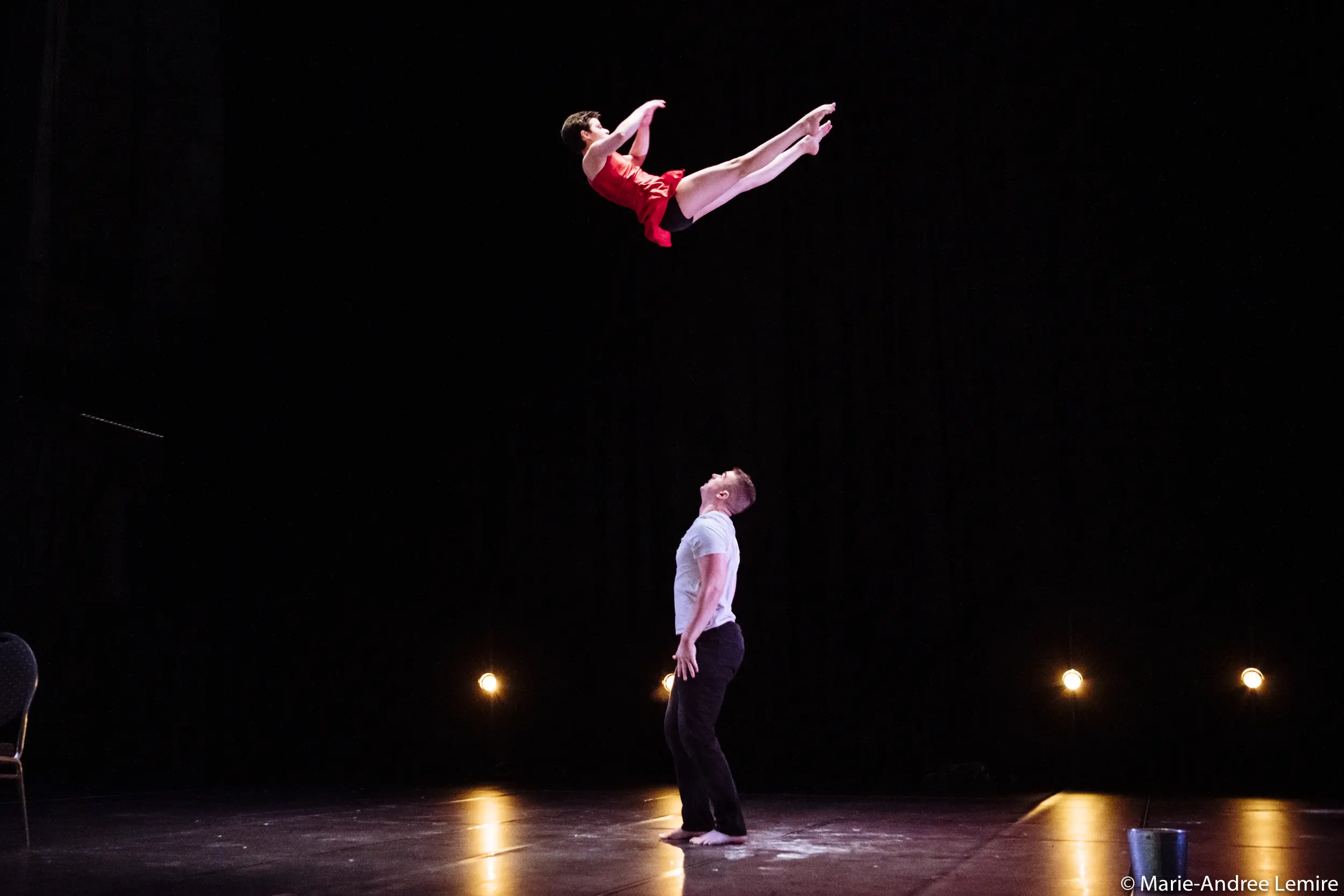 Un artiste masculin, Michael Carter, se tient sur scène et regarde en l'air tandis que l'acrobate féminine Frida Velasco, vêtue d'une tenue rouge, s'élance au-dessus de lui en plein vol, sur un fond sombre, les lumières de la scène éclairant la scène.
