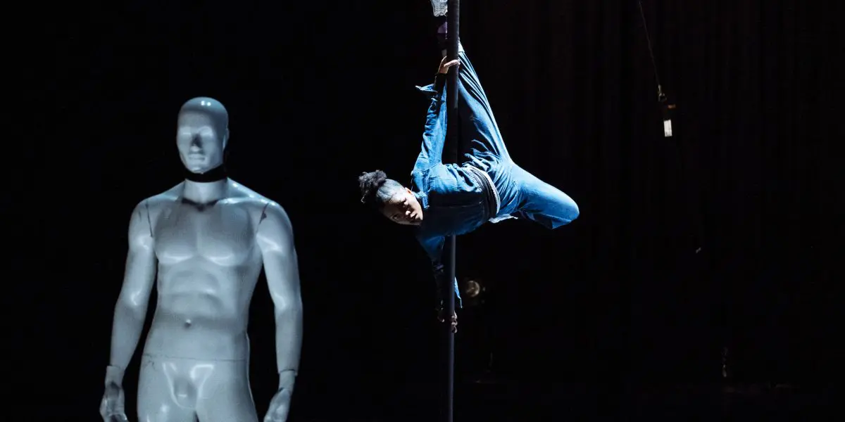 Naïka Aymon, vêtue de bleu, exécute un mouvement acrobatique sur un poteau vertical sur une scène sombre, tandis qu'un mannequin blanc à l'apparence humaine se tient au premier plan.
