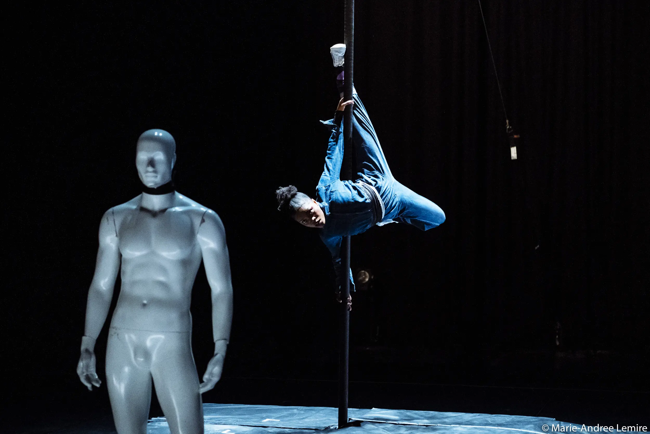 Naïka Aymon, vêtue de bleu, exécute un mouvement acrobatique sur un poteau vertical sur une scène sombre, tandis qu'un mannequin blanc à l'apparence humaine se tient au premier plan.