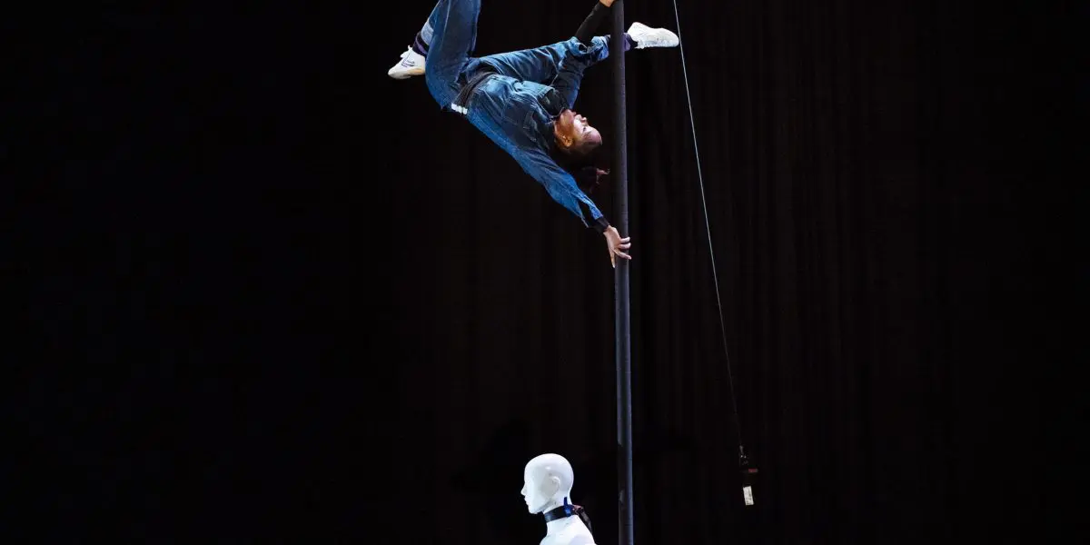Naïka Aymon, vêtue de bleu, est suspendue en l'air à un poteau vertical, tandis qu'un mannequin blanc se tient en bas sur une scène sombre.