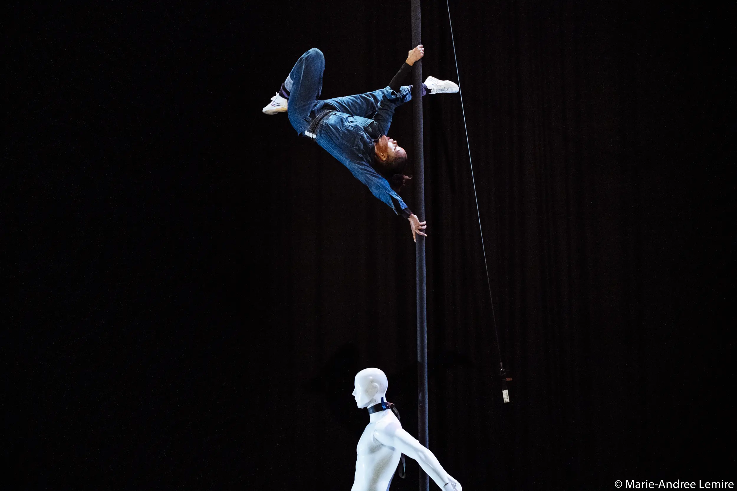 Naïka Aymon, vêtue de bleu, est suspendue en l'air à un poteau vertical, tandis qu'un mannequin blanc se tient en bas sur une scène sombre.