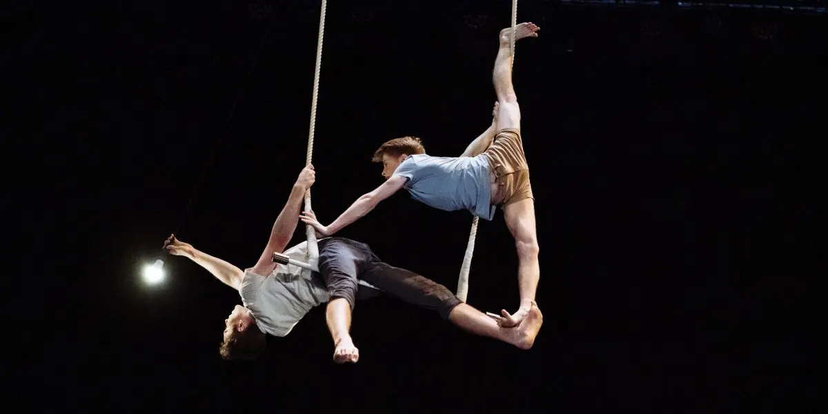 Deux artistes aériens, Maxime Charron et Corentin Lemaître Auger, se tiennent en équilibre sur des cordes suspendues, l'un tendant une jambe en hauteur, l'autre s'étirant tout en saisissant une corde, le tout sur un fond noir dramatique.