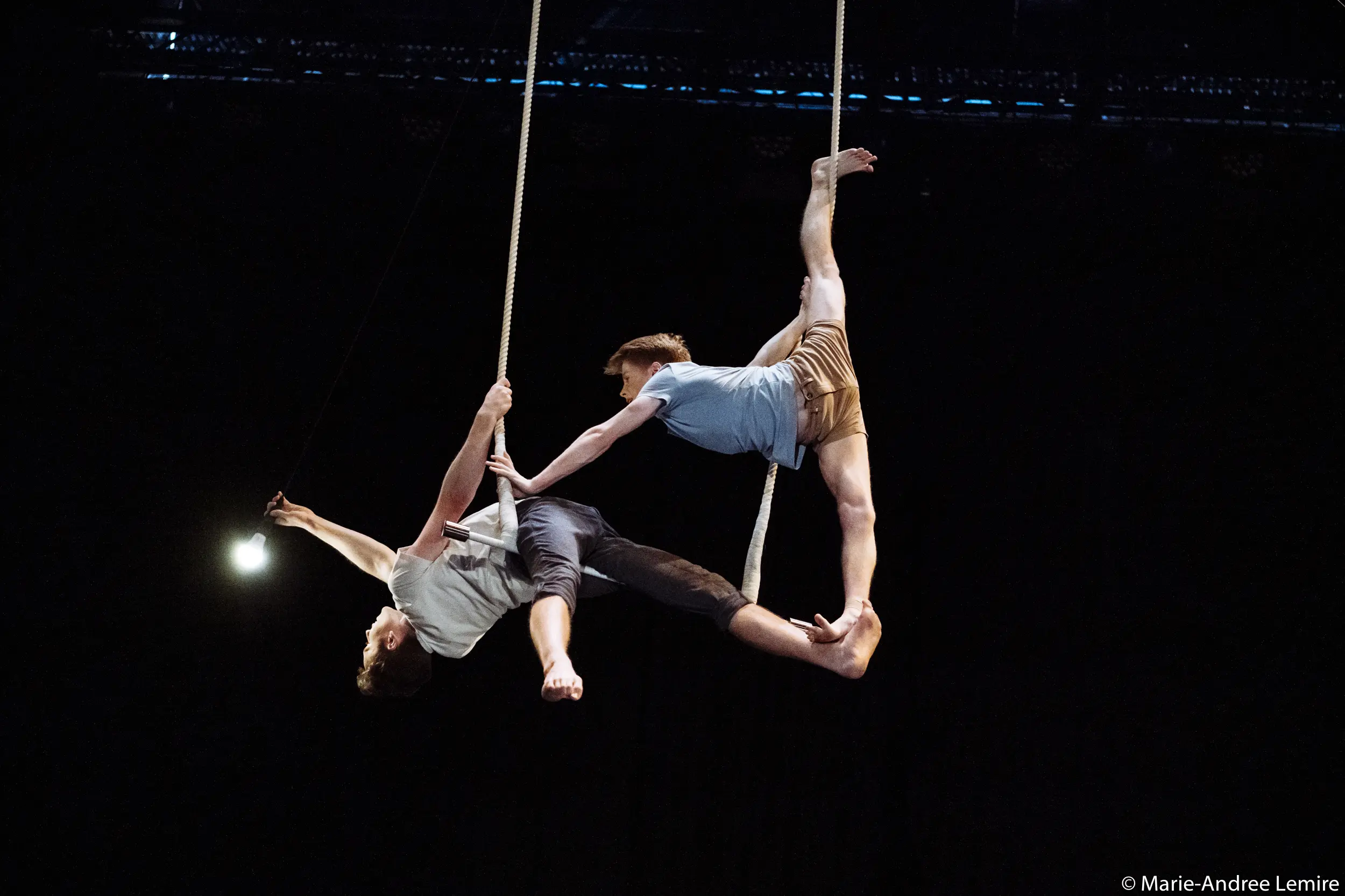 Deux artistes aériens, Maxime Charron et Corentin Lemaître Auger, se tiennent en équilibre sur des cordes suspendues, l'un tendant une jambe en hauteur, l'autre s'étirant tout en saisissant une corde, le tout sur un fond noir dramatique.