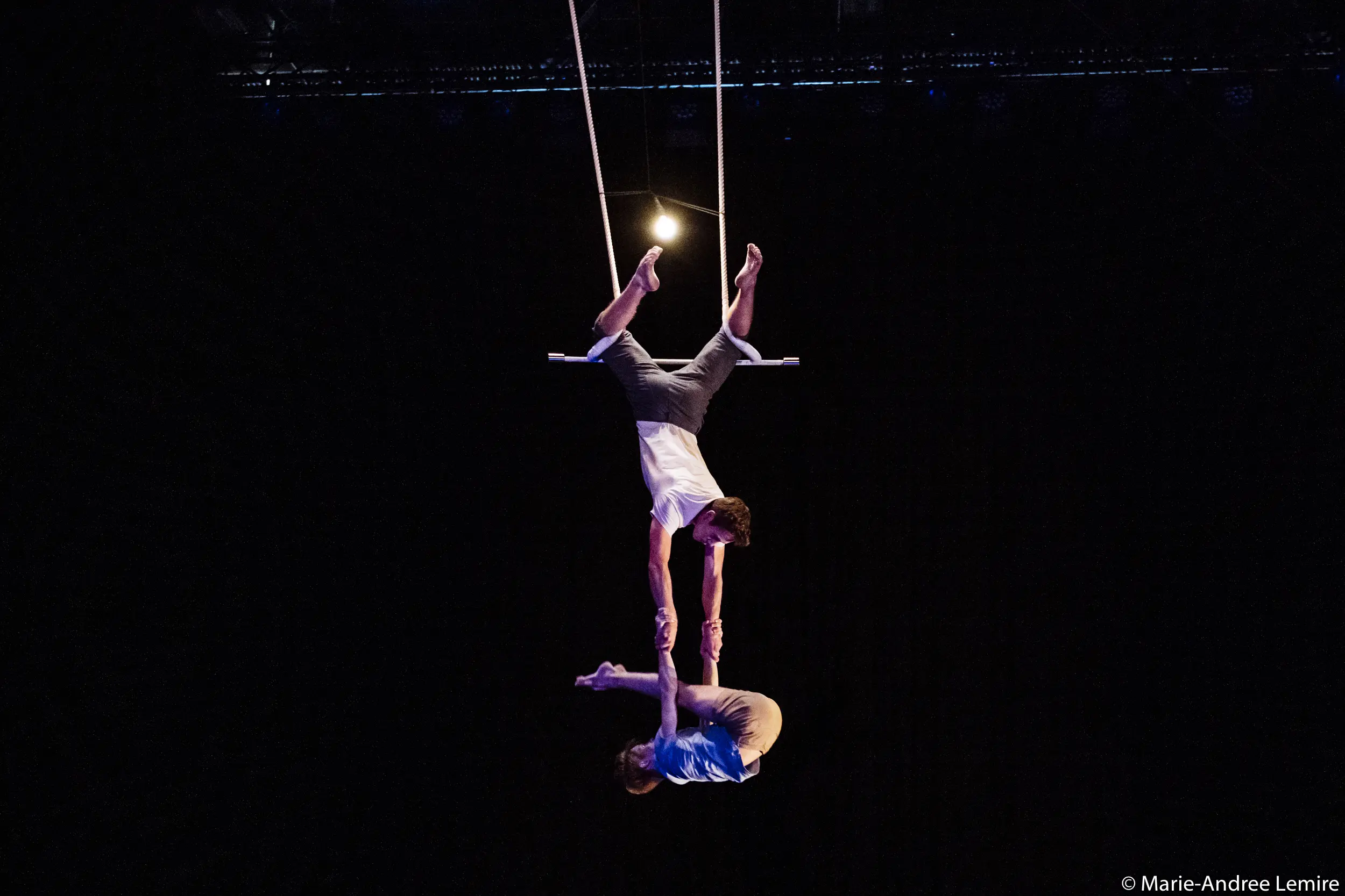 Deux acrobates, Maxime Charron et Corentin Lemaître Auger, évoluent sur un trapèze - l'un suspendu à l'envers par les jambes, l'autre agrippé aux bras de l'autre qui se balance en dessous d'eux - sur un fond sombre.