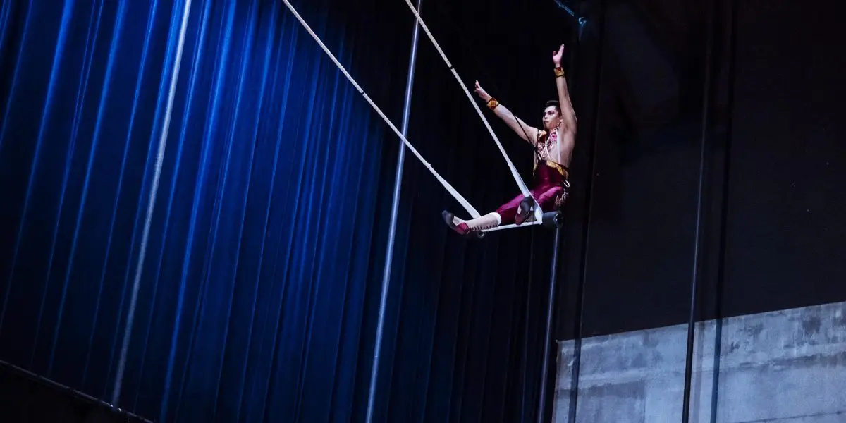 Gabriel Olmos Cruz, un artiste de cirque vêtu d'un costume rouge, se produit sur un trapèze, suspendu très haut au-dessus du sol, un bras levé, sur fond de rideaux bleus et de mur en béton.