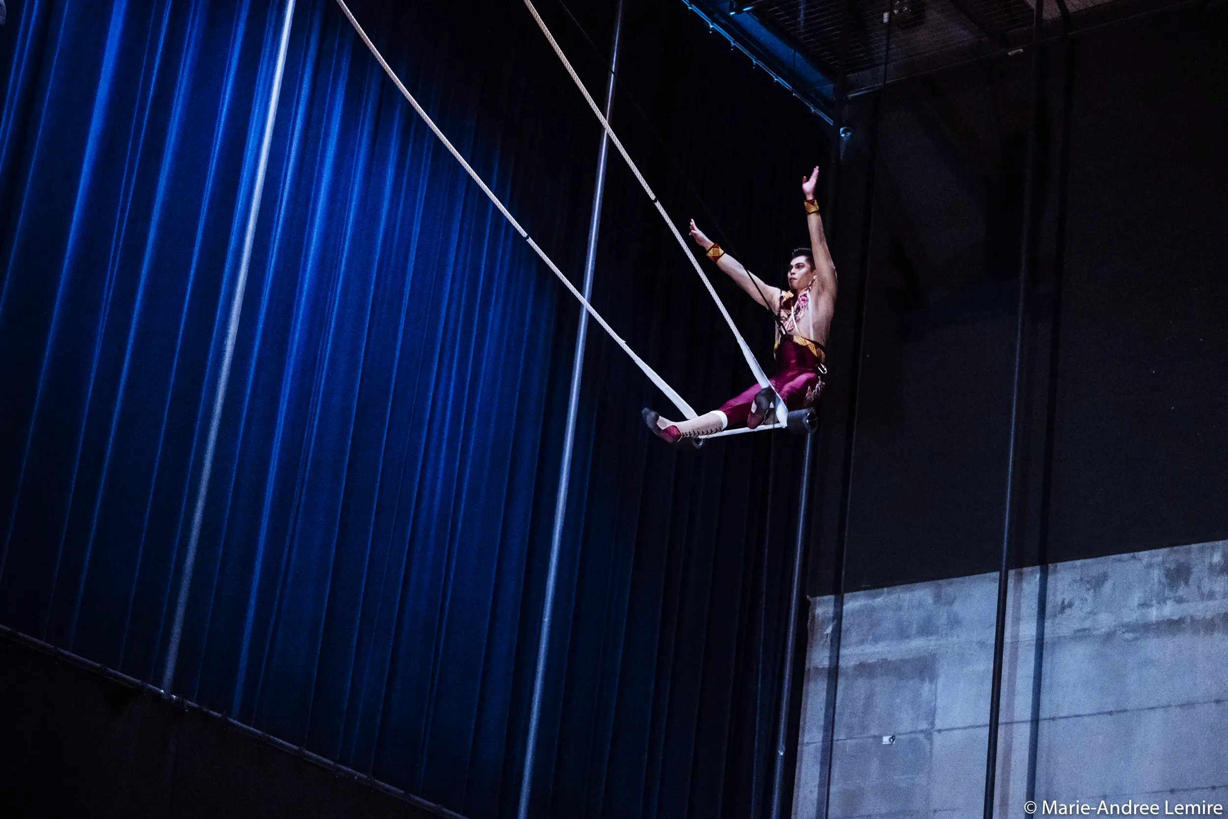 Gabriel Olmos Cruz, un artiste de cirque vêtu d'un costume rouge, se produit sur un trapèze, suspendu très haut au-dessus du sol, un bras levé, sur fond de rideaux bleus et de mur en béton.