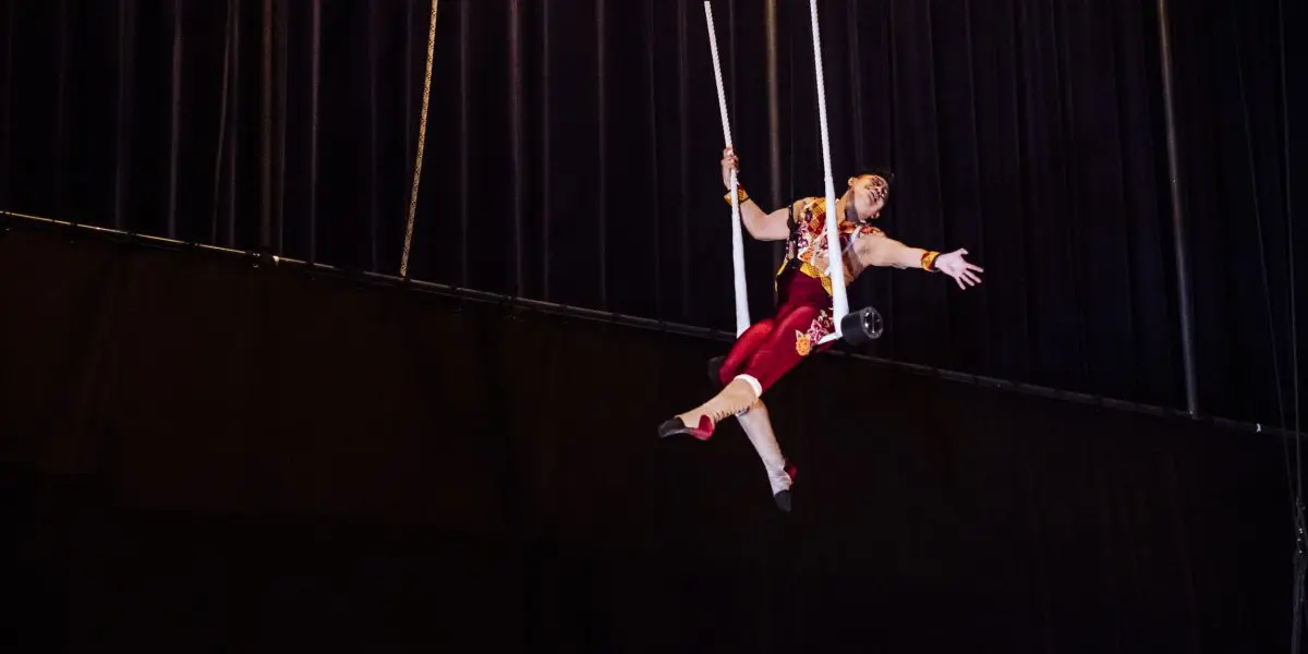 Un artiste de cirque en costume rouge et or est suspendu en l'air sur un trapèze, étendant gracieusement un bras, avec des rideaux sombres en arrière-plan, rappelant l'art de Gabriel Olmos Cruz. © Marie-Andree Lemire.