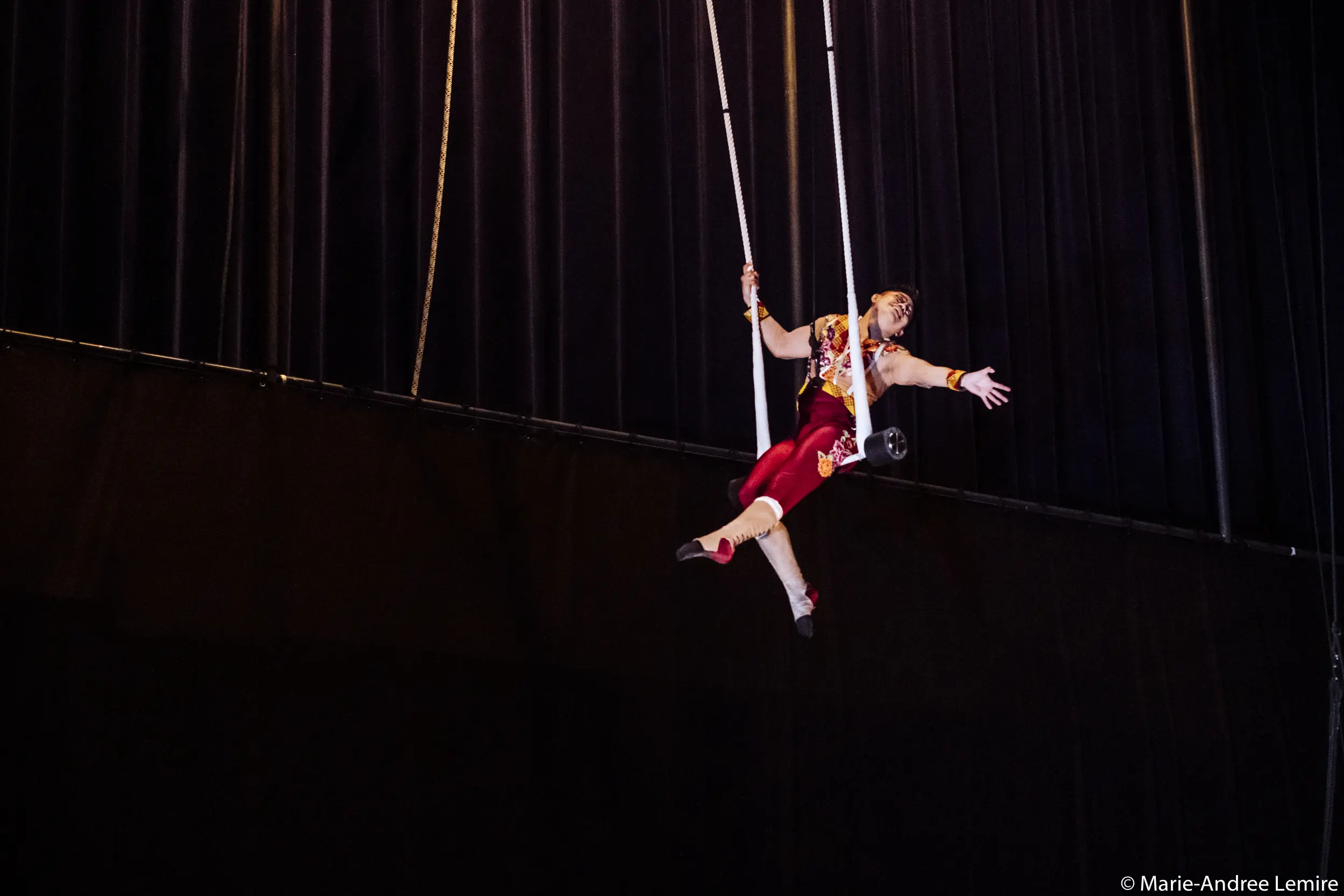 Un artiste de cirque en costume rouge et or est suspendu en l'air sur un trapèze, étendant gracieusement un bras, avec des rideaux sombres en arrière-plan, rappelant l'art de Gabriel Olmos Cruz. © Marie-Andree Lemire.