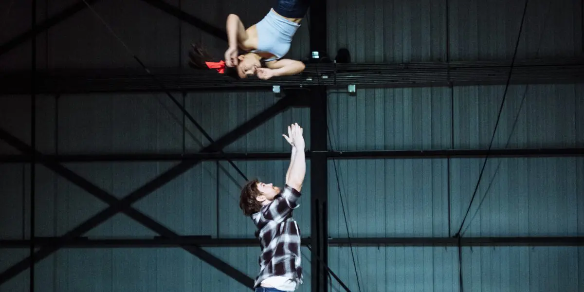 Une personne vêtue d'une chemise à carreaux se tient debout sur un tapis, les bras levés, et attrape Ashley Youren, qui est en train de voler et de se retourner au-dessus d'eux, dans une grande arène intérieure.