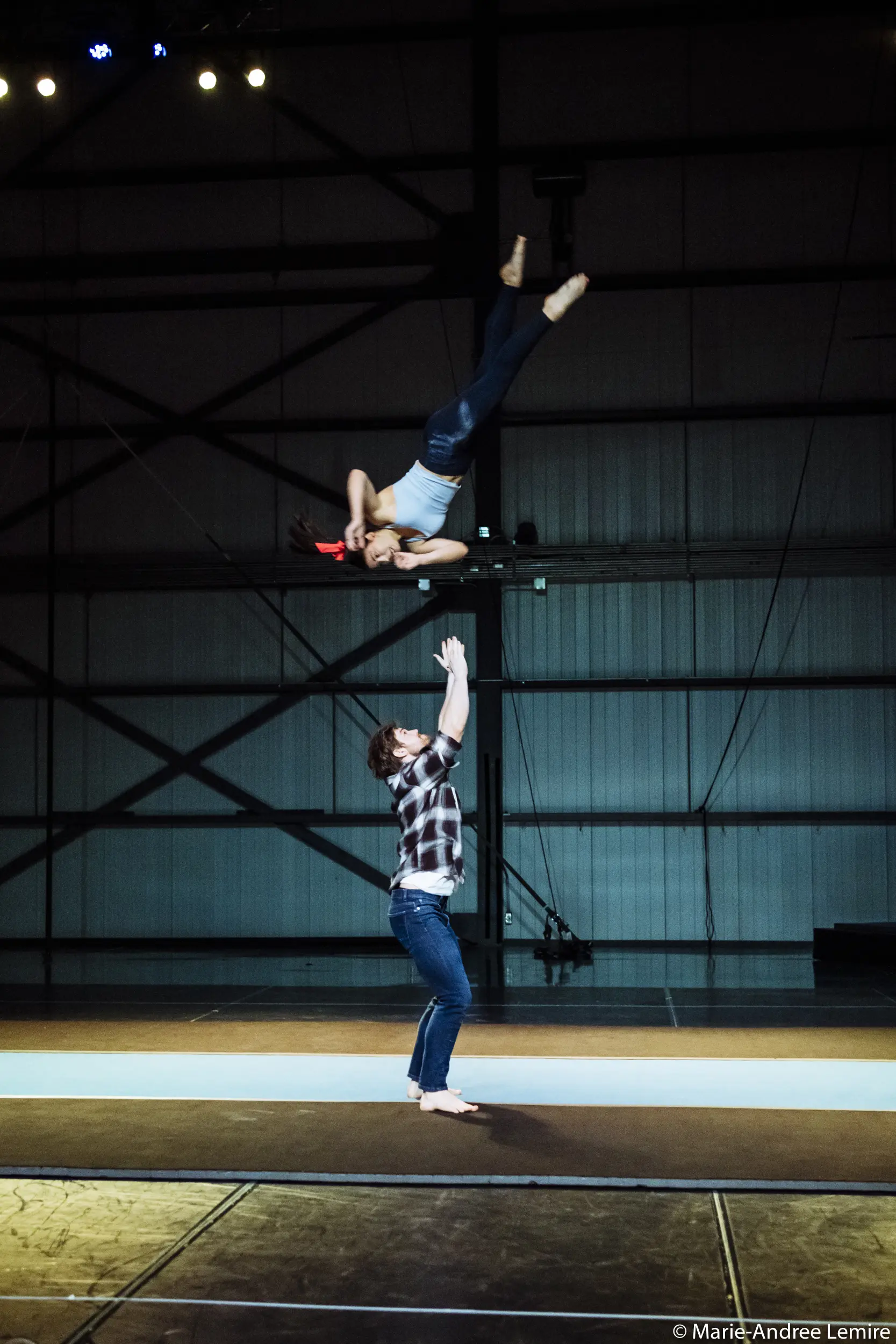 Une personne vêtue d'une chemise à carreaux se tient debout sur un tapis, les bras levés, et attrape Ashley Youren, qui est en train de voler et de se retourner au-dessus d'eux, dans une grande arène intérieure.