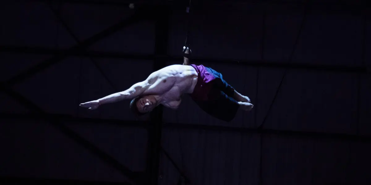 Samuel Donald Nash, un voltigeur masculin aux bras tendus, exécute un mouvement acrobatique en étant suspendu par une sangle dans un décor intérieur sombre éclairé par un projecteur.