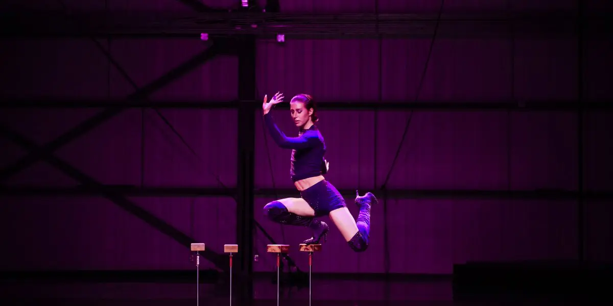Un artiste en tenue de sport noire, rappelant le style dynamique de Yann Lortie, saute gracieusement en l'air sur la scène, bras levés et genoux pliés, sous un éclairage violet. De petites plates-formes en contrebas évoquent un numéro de cirque ou d'acrobatie.