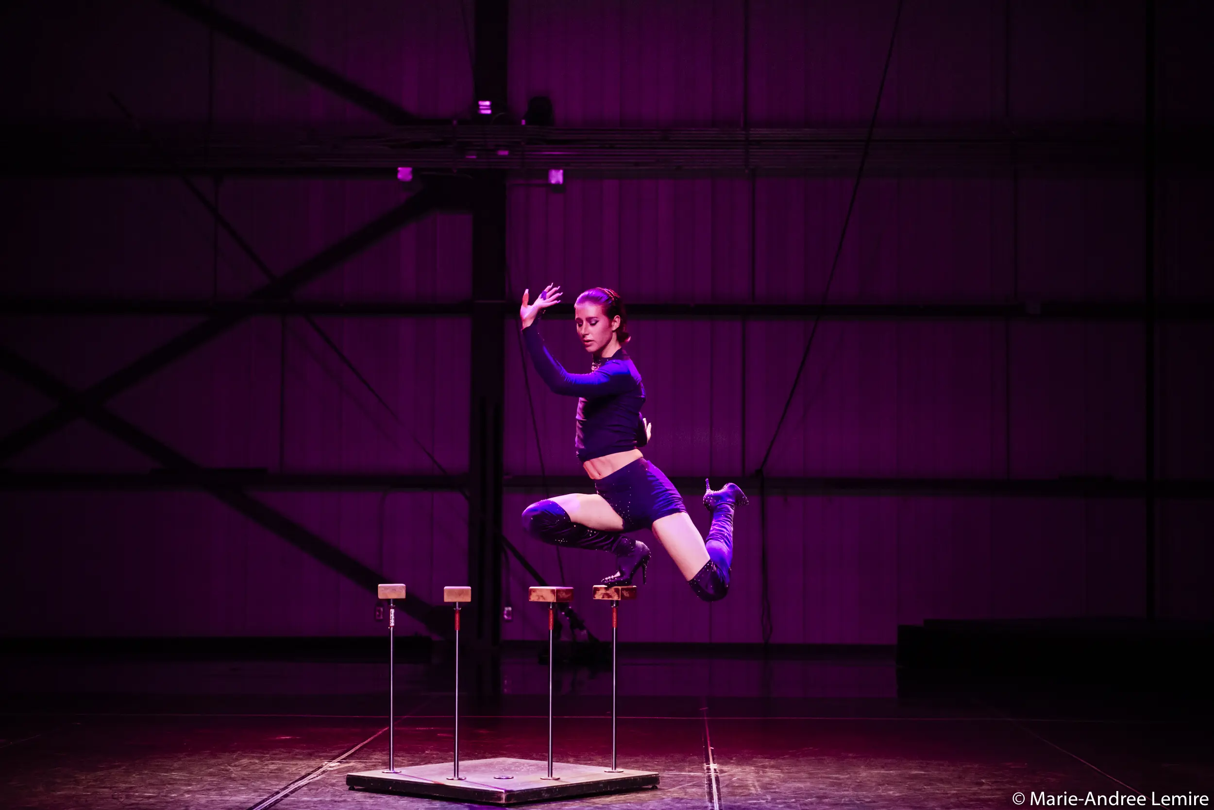 Un artiste en tenue de sport noire, rappelant le style dynamique de Yann Lortie, saute gracieusement en l'air sur la scène, bras levés et genoux pliés, sous un éclairage violet. De petites plates-formes en contrebas évoquent un numéro de cirque ou d'acrobatie.