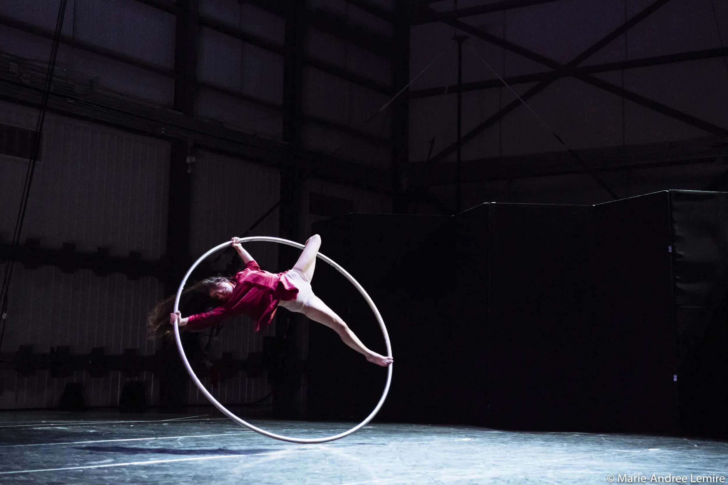 Une artiste en tenue rouge se suspend gracieusement à un grand cerceau aérien sur une scène faiblement éclairée avec des murs industriels en arrière-plan - un numéro inoubliable d'Andrea Martinez Aguilar.