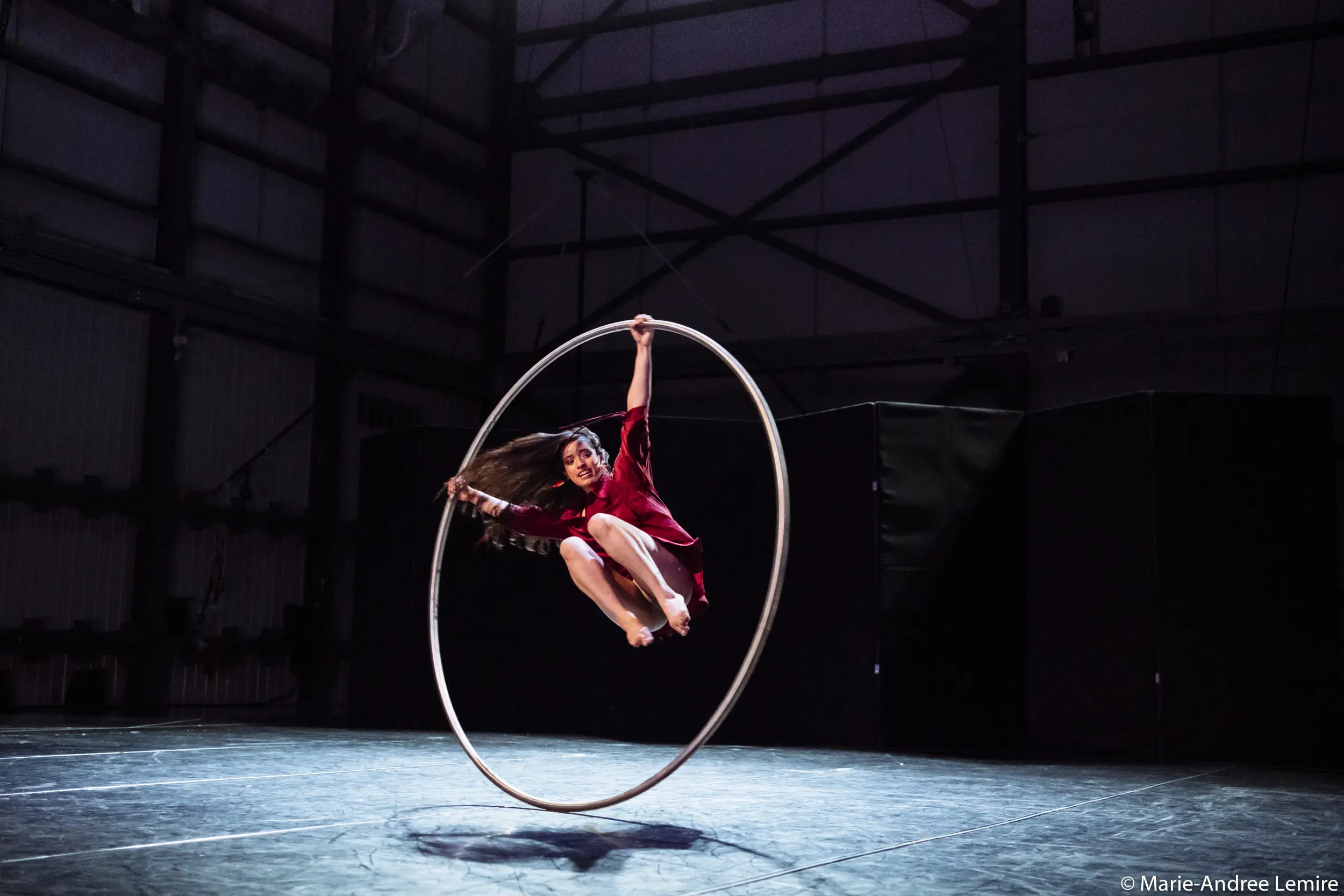 Andrea Martinez Aguilar, dans une tenue rouge frappante, se tient gracieusement en équilibre à l'intérieur d'un grand cerceau métallique, en pleine représentation, sur une scène faiblement éclairée avec des murs de style industriel en arrière-plan.