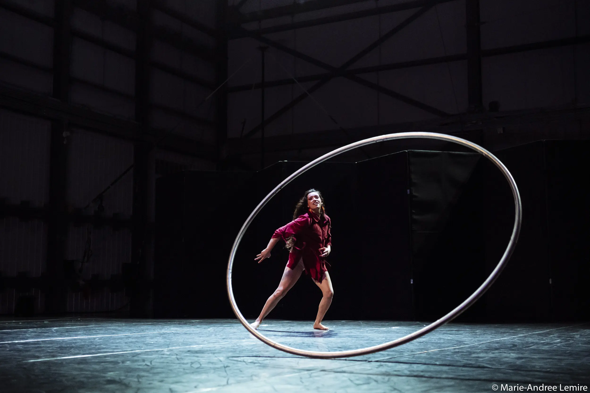 Andrea Martinez Aguilar, vêtue d'une tenue rouge éclatante, se tient sur une scène faiblement éclairée, se balançant gracieusement dans un grand cerceau métallique au milieu d'ombres dramatiques et d'un arrière-plan industriel.
