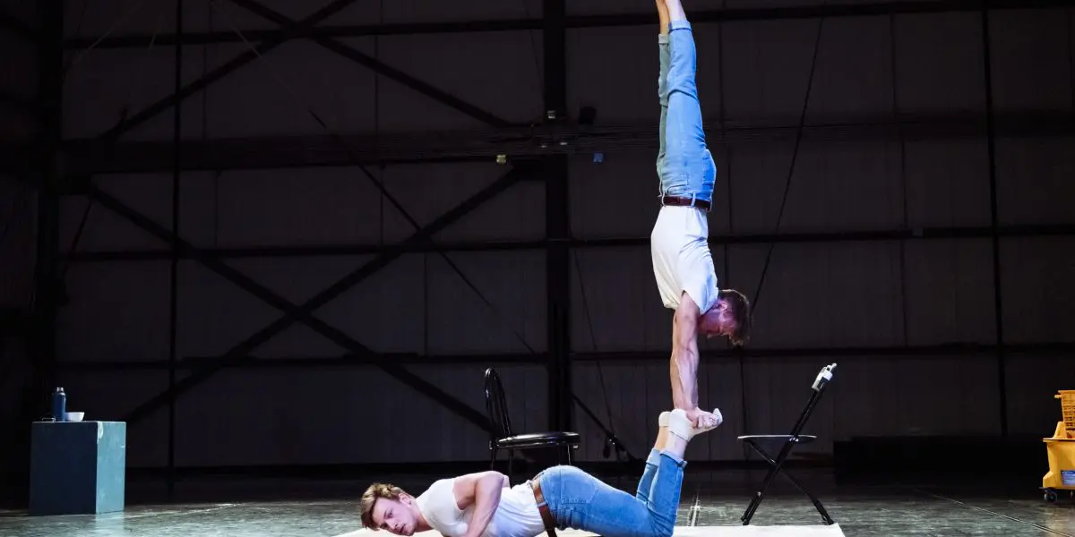 Deux artistes en jeans et chemise blanche prennent une pose acrobatique : l'une est allongée sur le ventre, les jambes pliées, tandis que l'autre se tient en équilibre sur ses pieds, la tête en bas, en se tenant les chevilles - une scène saisissante qui rappelle le style dynamique de Benjamin Wolgers.