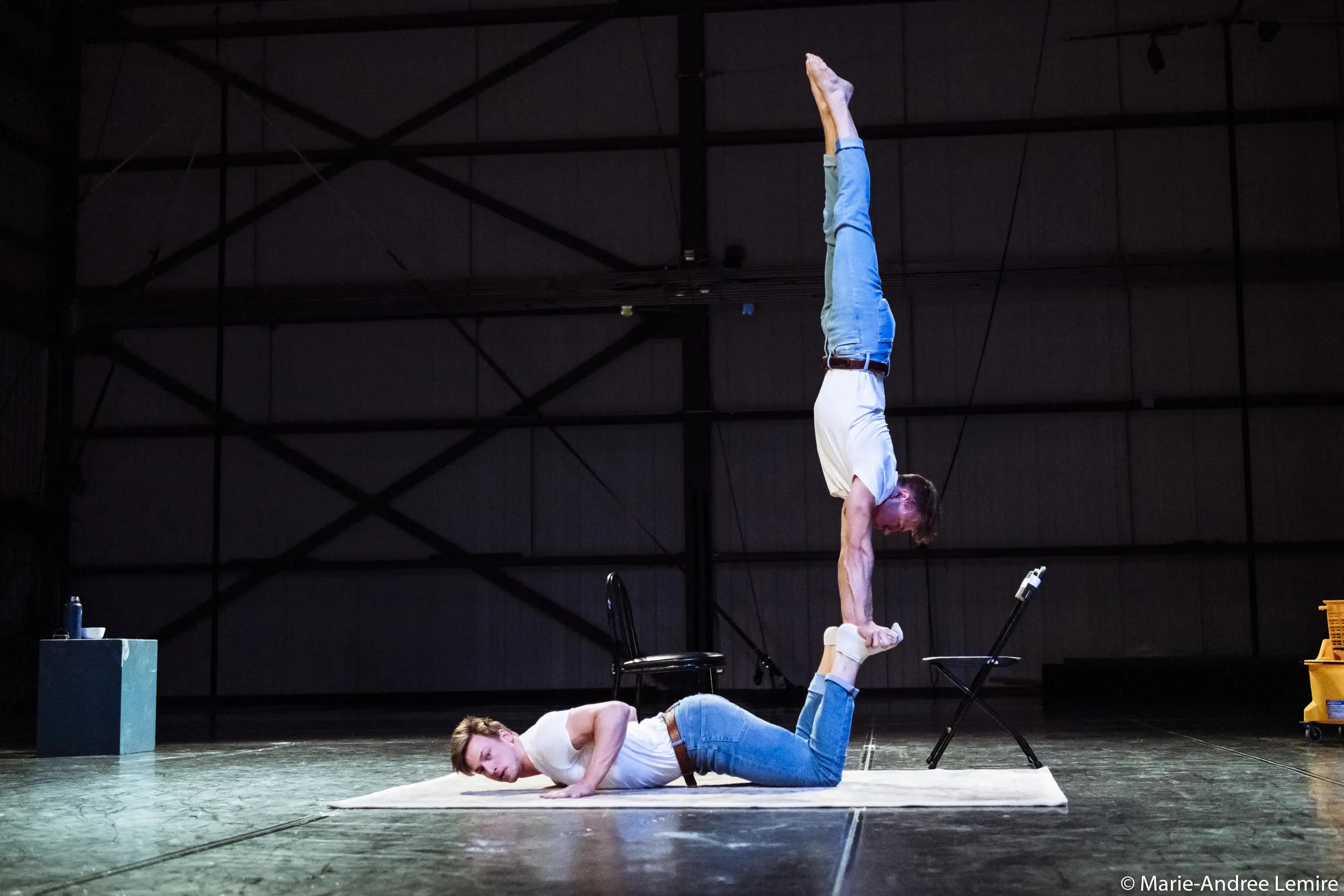 Deux artistes en jeans et chemise blanche prennent une pose acrobatique : l'une est allongée sur le ventre, les jambes pliées, tandis que l'autre se tient en équilibre sur ses pieds, la tête en bas, en se tenant les chevilles - une scène saisissante qui rappelle le style dynamique de Benjamin Wolgers.