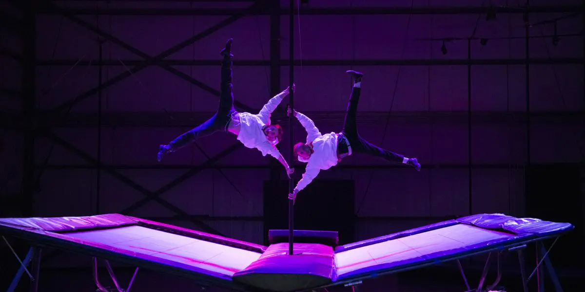 Deux acrobates, dont Mathys Guyon Matte, vêtus de chemises blanches et de pantalons sombres, réalisent un équilibre synchronisé sur une perche, en se balançant dans les airs au-dessus de trampolines, sous un éclairage dramatique de couleur violette.