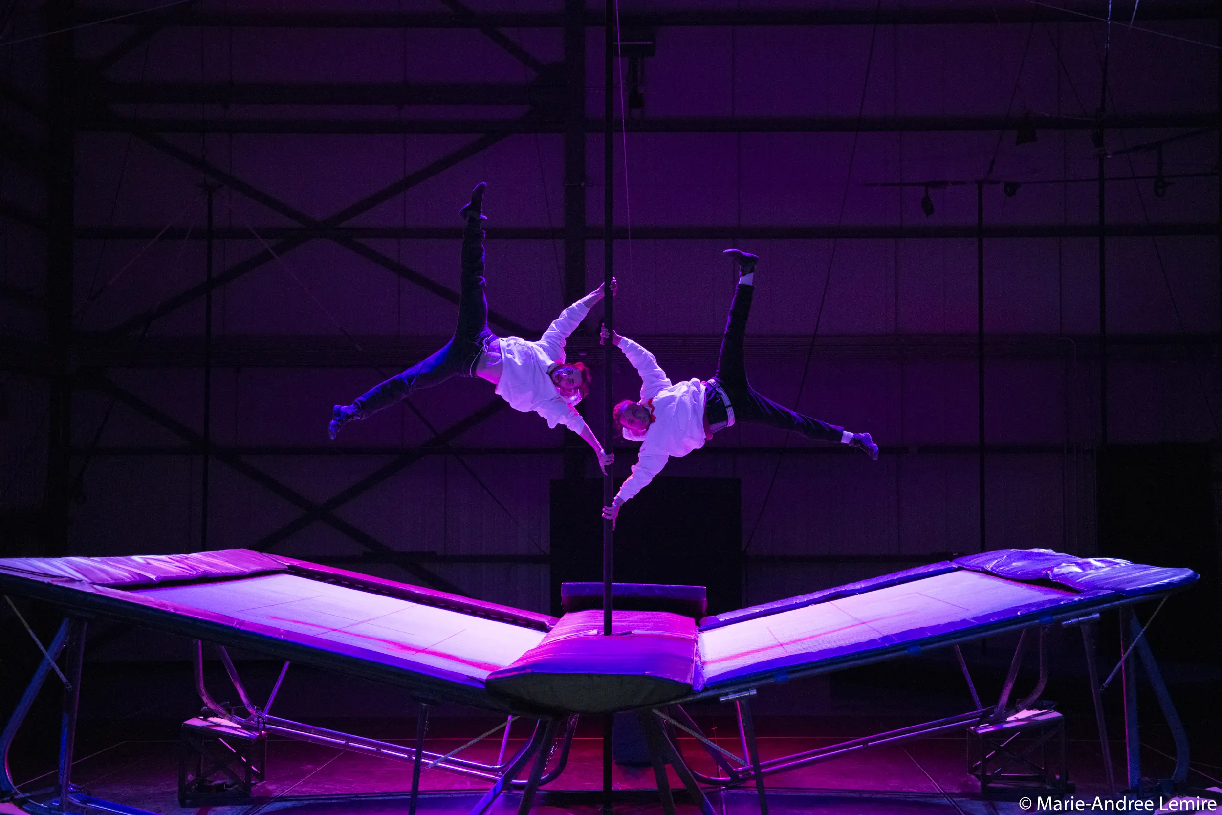 Deux acrobates, dont Mathys Guyon Matte, vêtus de chemises blanches et de pantalons sombres, réalisent un équilibre synchronisé sur une perche, en se balançant dans les airs au-dessus de trampolines, sous un éclairage dramatique de couleur violette.