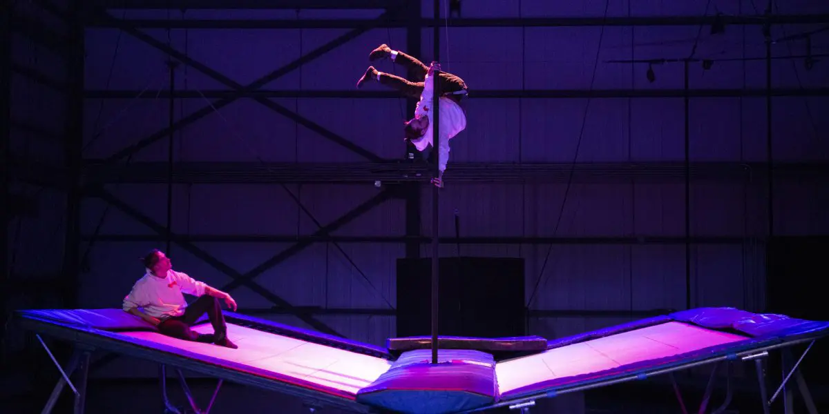 Deux acrobates, dont Mathys Guyon Matte, évoluent sur des trampolines sous un éclairage de scène violet ; l'un est assis sur le trampoline de gauche tandis que l'autre prend une pose inversée en haut d'un poteau vertical au centre.