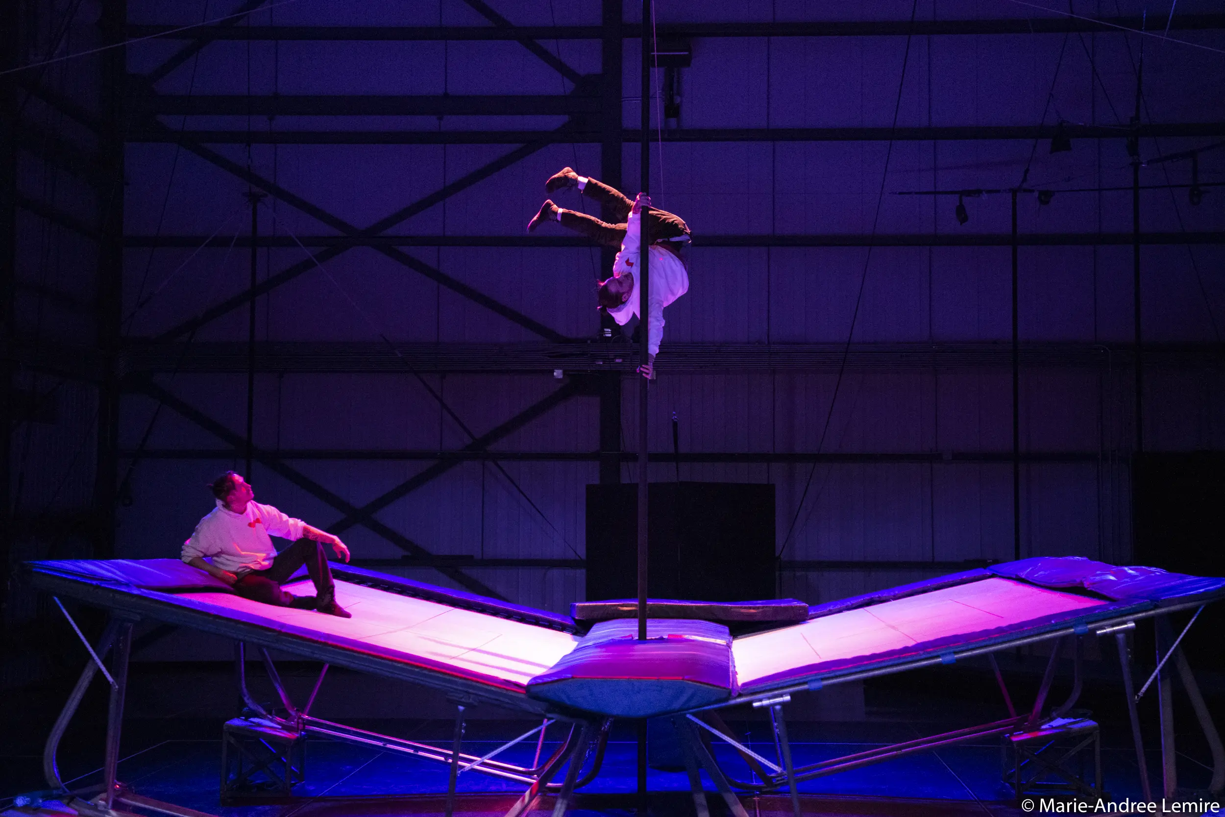 Deux acrobates, dont Mathys Guyon Matte, évoluent sur des trampolines sous un éclairage de scène violet ; l'un est assis sur le trampoline de gauche tandis que l'autre prend une pose inversée en haut d'un poteau vertical au centre.