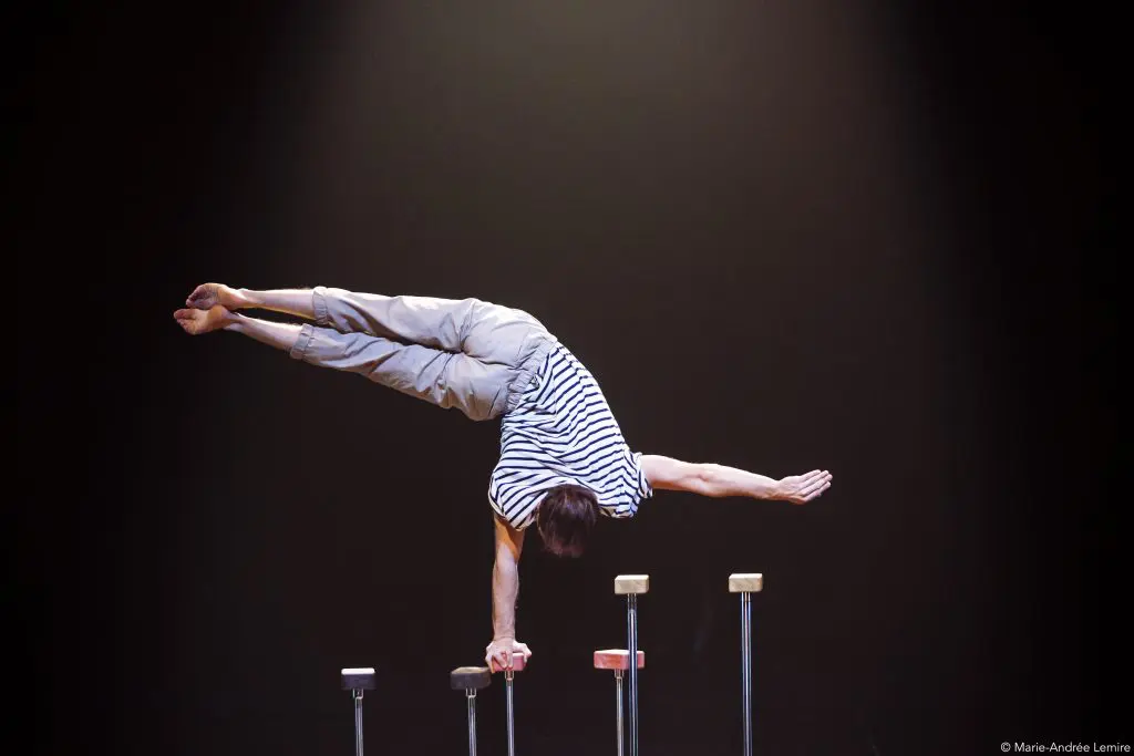 Carl Löwenborg, vêtu d'une chemise rayée et d'un pantalon clair, exécute un exercice d'équilibre sur un bras, les jambes tendues latéralement et l'autre bras tendu, sur une scène sombre.