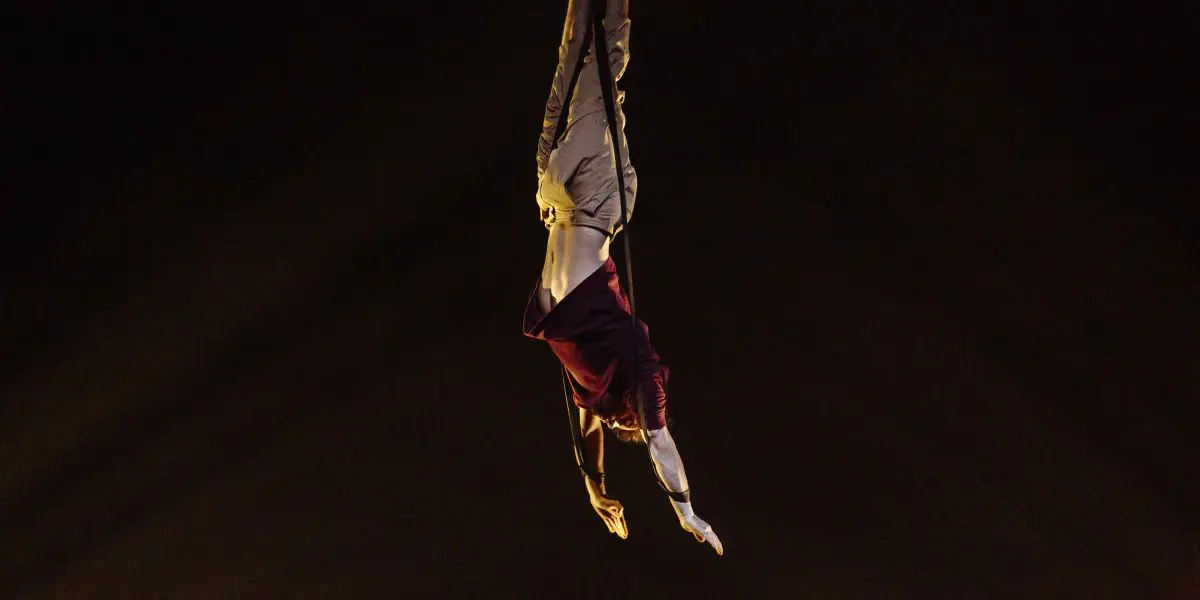 Un acrobate vêtu d'une chemise marron et d'un pantalon beige est suspendu à l'envers dans les airs, les bras étendus, sur un fond sombre, dramatiquement éclairé par le bas - une capture de l'art d'Eduardo De Azevedo Grillo.
