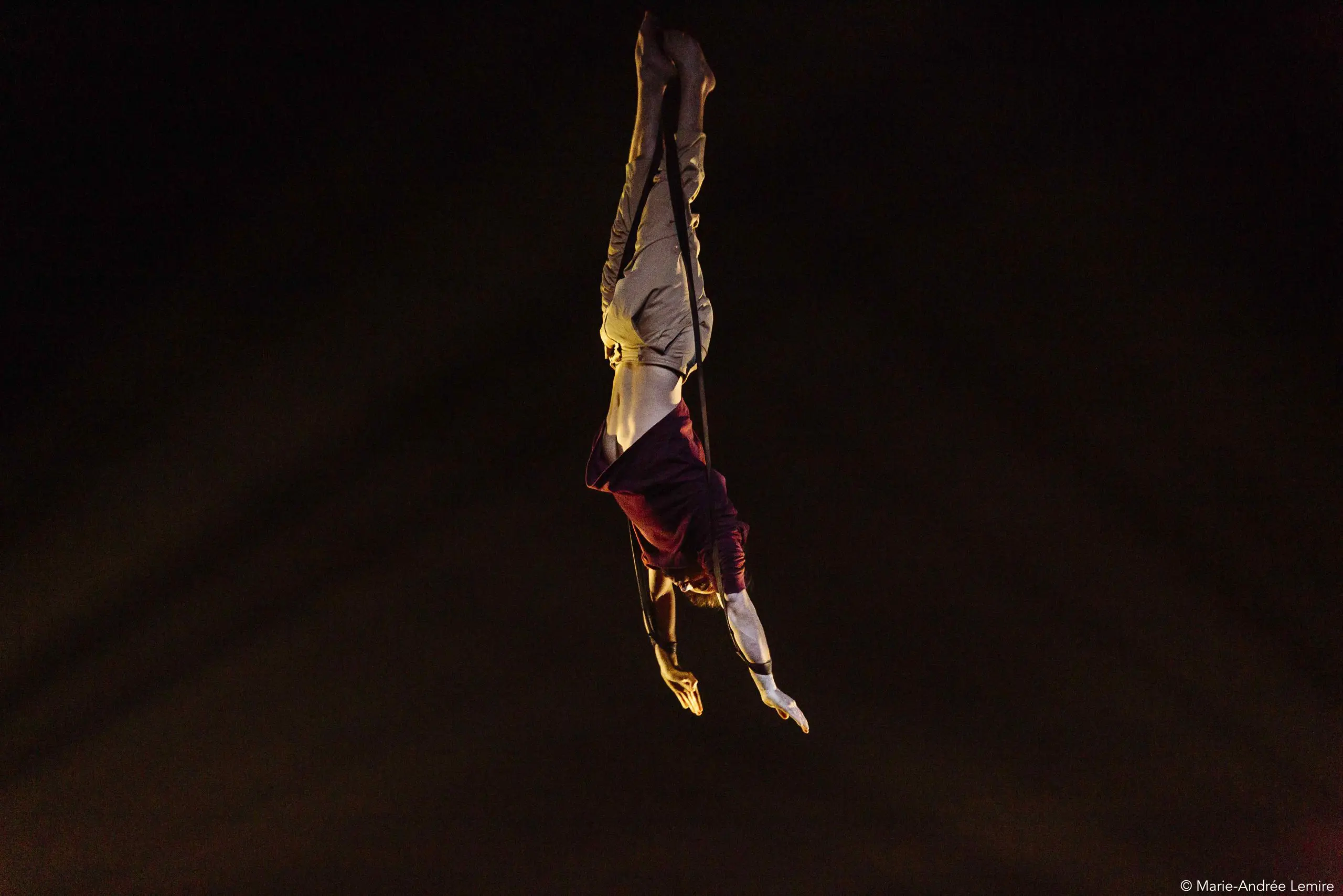 Un acrobate vêtu d'une chemise marron et d'un pantalon beige est suspendu à l'envers dans les airs, les bras étendus, sur un fond sombre, dramatiquement éclairé par le bas - une capture de l'art d'Eduardo De Azevedo Grillo.
