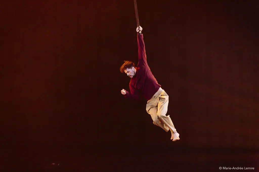 Un artiste, Eduardo De Azevedo Grillo, vêtu d'une chemise marron et d'un pantalon beige, est suspendu d'une main à une sangle, dans les airs, sur un fond sombre. Le corps de la personne est incliné et ses pieds nus sont visibles.