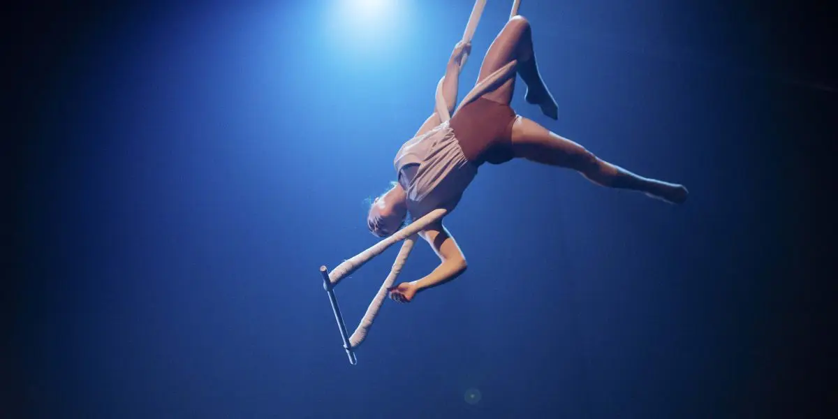 L'acrobate aérienne Daphnée Sergerie évolue sur des sangles aériennes blanches sous un projecteur, gracieusement suspendue dans les airs sur un fond sombre et éclairé en bleu.