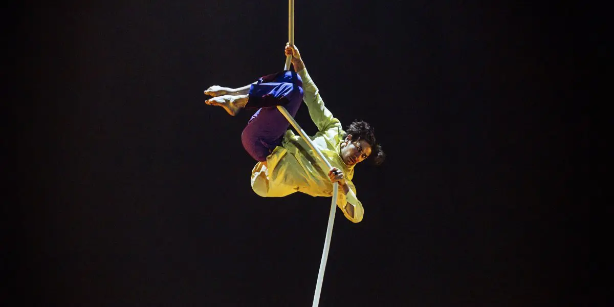 L'artiste de cirque Dylan Ingwersen, vêtu d'une chemise jaune et d'un pantalon bleu, est suspendu la tête en bas tout en saisissant une corde aérienne blanche sur un fond sombre.