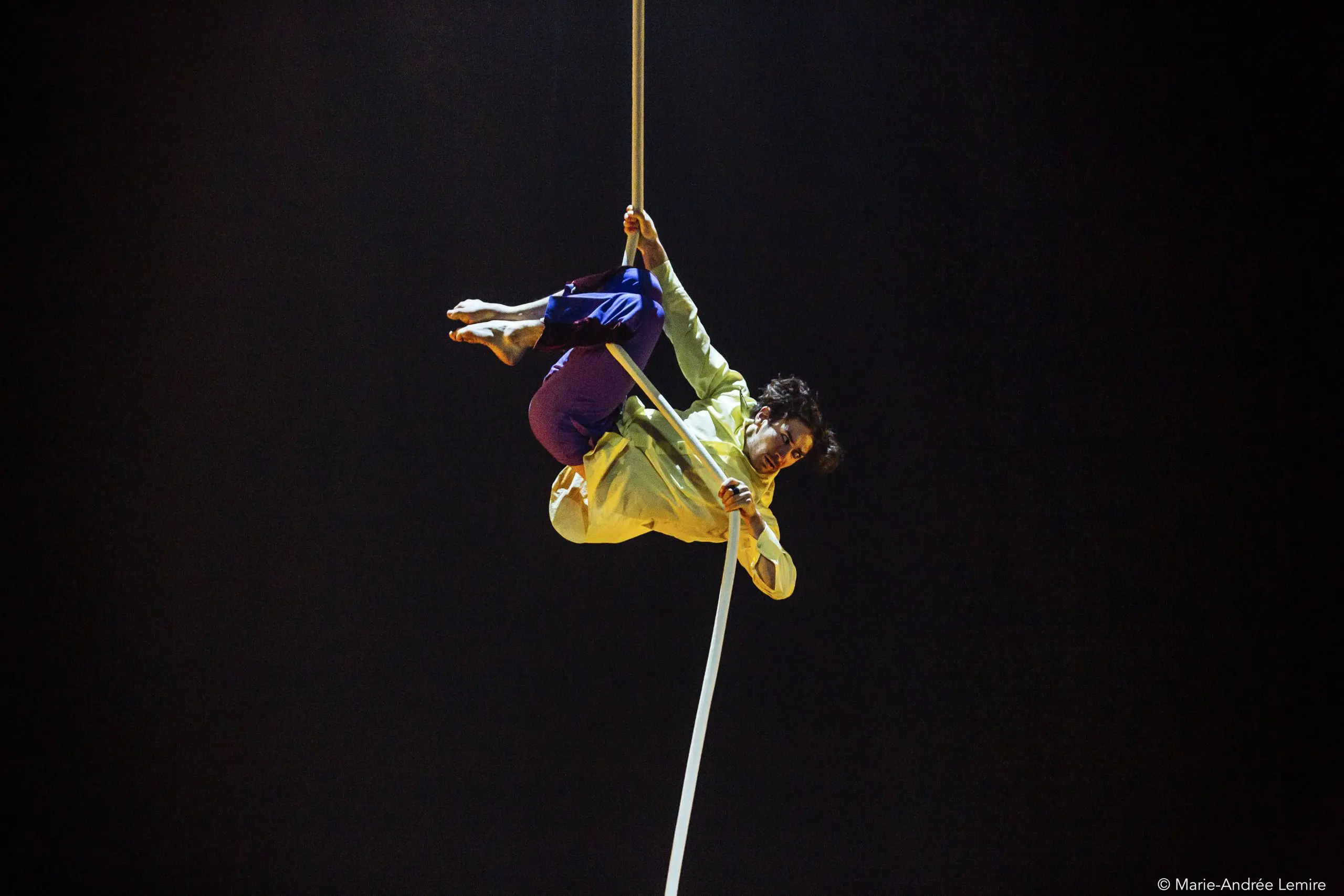 L'artiste de cirque Dylan Ingwersen, vêtu d'une chemise jaune et d'un pantalon bleu, est suspendu la tête en bas tout en saisissant une corde aérienne blanche sur un fond sombre.