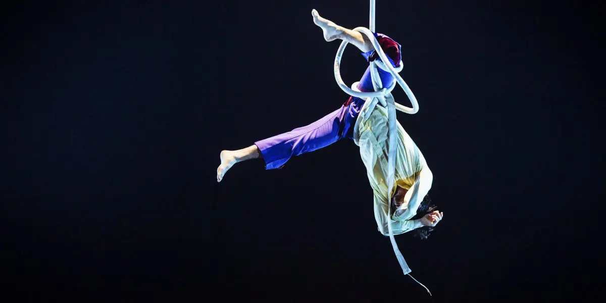 Deux artistes aériens, dont Dylan Ingwersen, sont suspendus à l'envers sur un cerceau blanc, avec un éclairage de scène dramatique et un arrière-plan sombre mettant en valeur leur pose acrobatique.