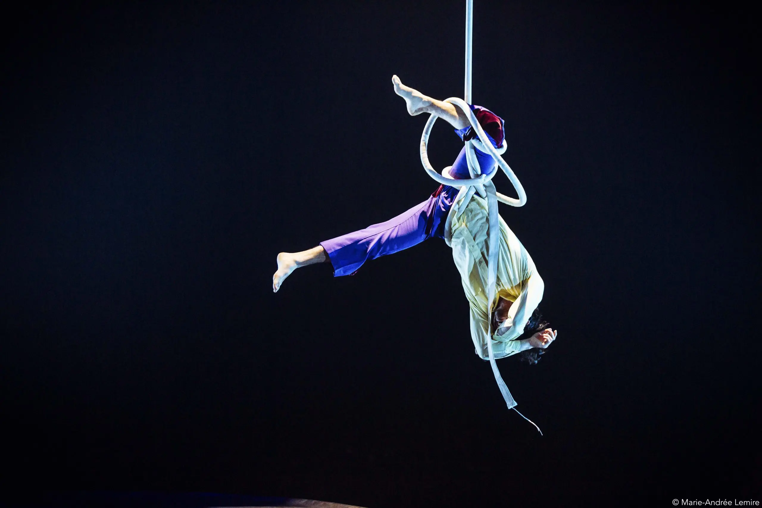 Deux artistes aériens, dont Dylan Ingwersen, sont suspendus à l'envers sur un cerceau blanc, avec un éclairage de scène dramatique et un arrière-plan sombre mettant en valeur leur pose acrobatique.