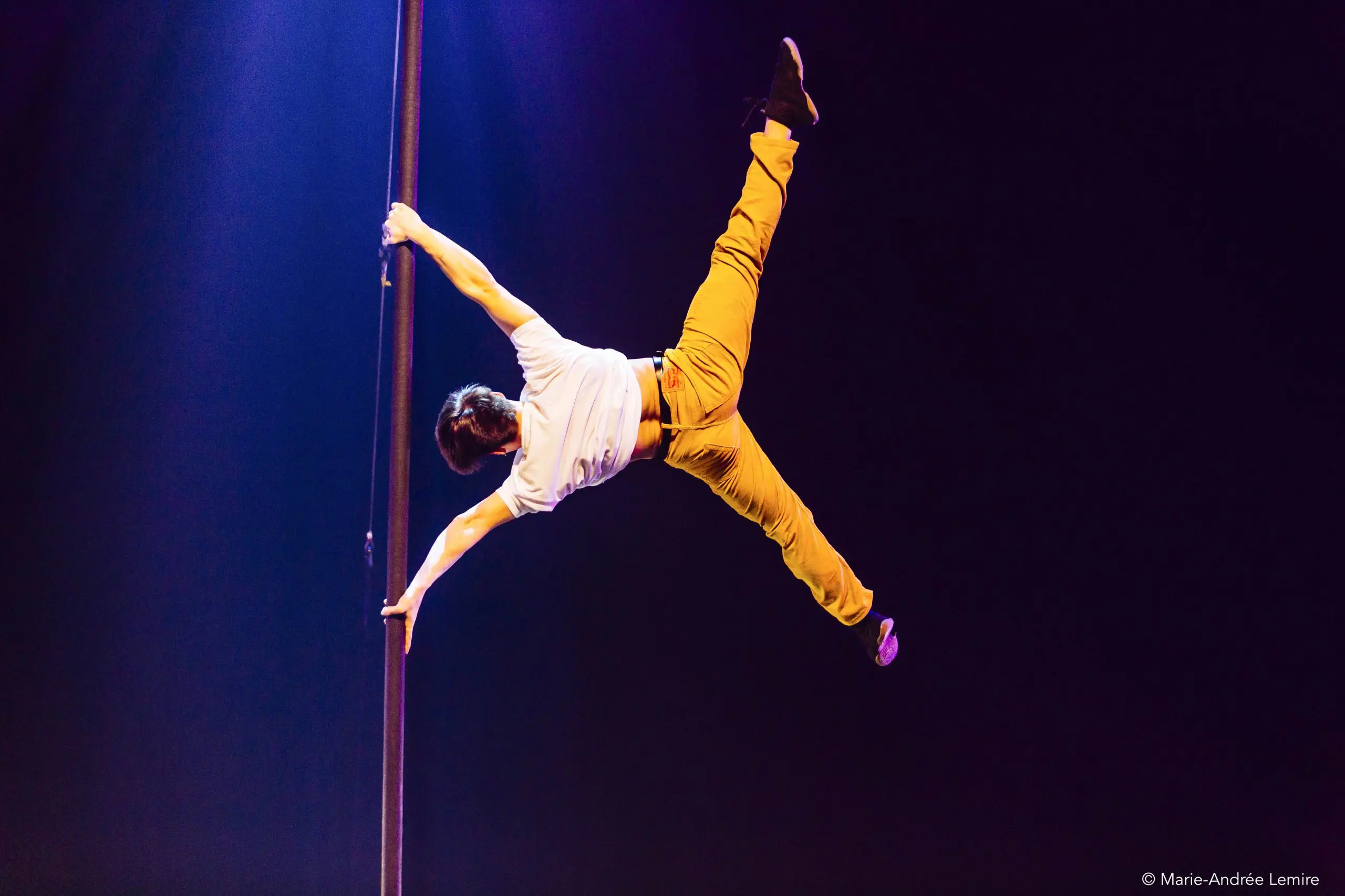 Antoine Brabant effectue une acrobatie à la perche, en saisissant une perche verticale d'une main tout en étendant son corps à l'horizontale. Il est vêtu d'une chemise blanche et d'un pantalon jaune sur fond sombre.