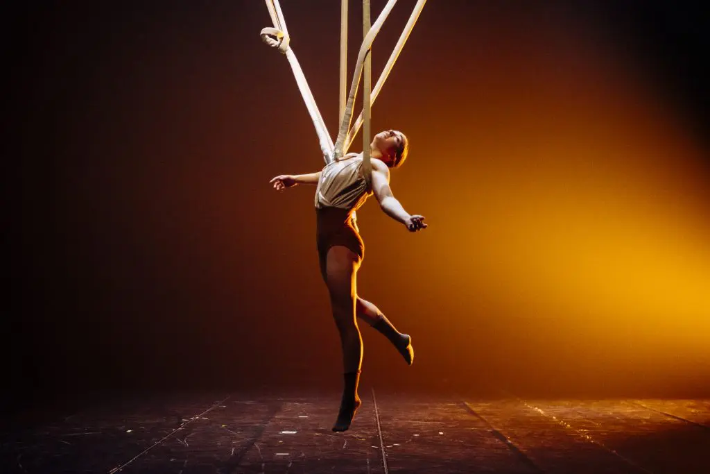 La performeuse aérienne Daphnée Sergerie est suspendue en l'air sur la scène, enveloppée dans un long tissu blanc, avec un bras tendu et un éclairage jaune-orange dramatique qui illumine l'arrière-plan.